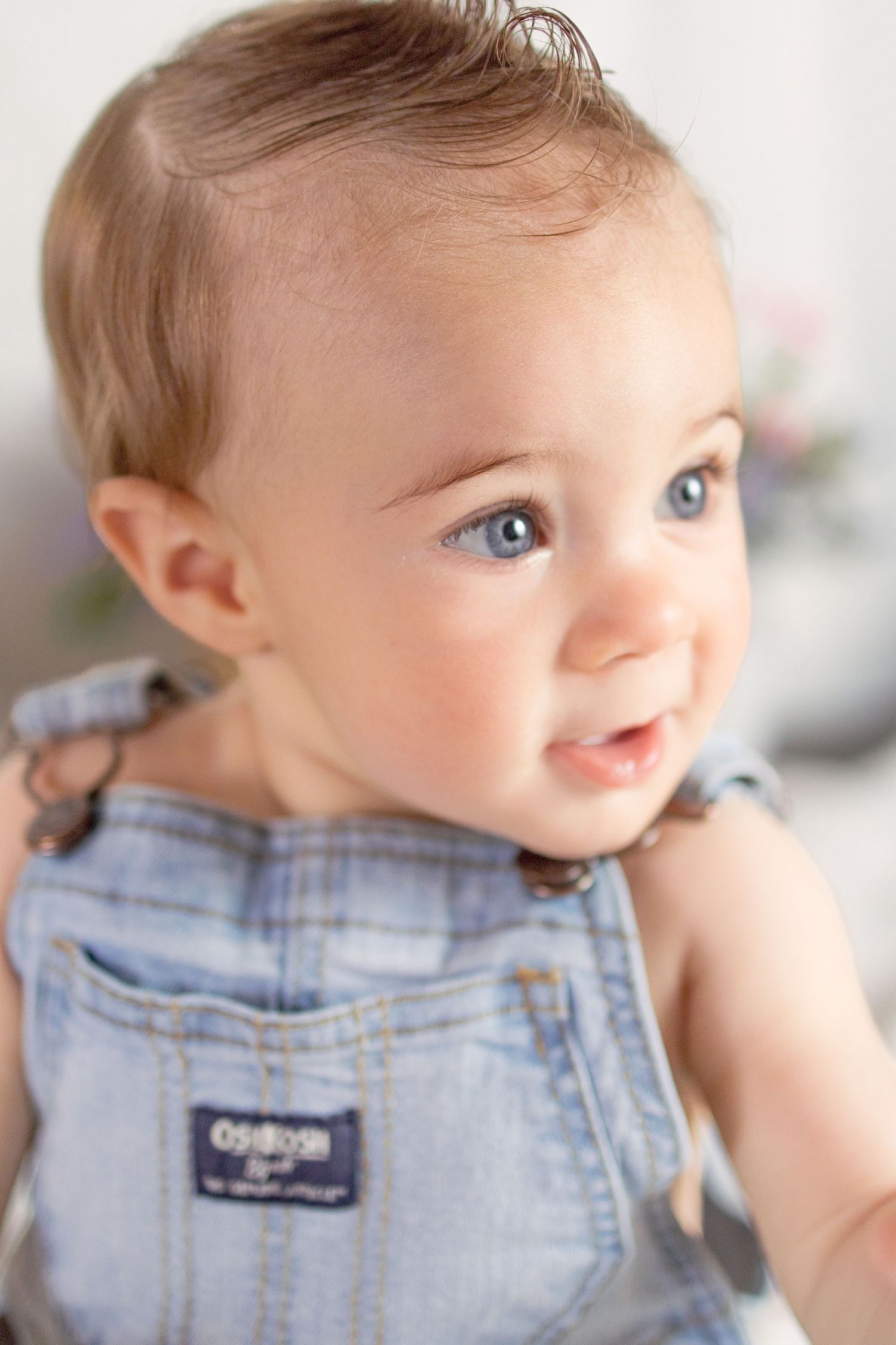 Adorable baby with blue eyes wearing denim OshKosh overalls smiling softly in a bright portrait photo.