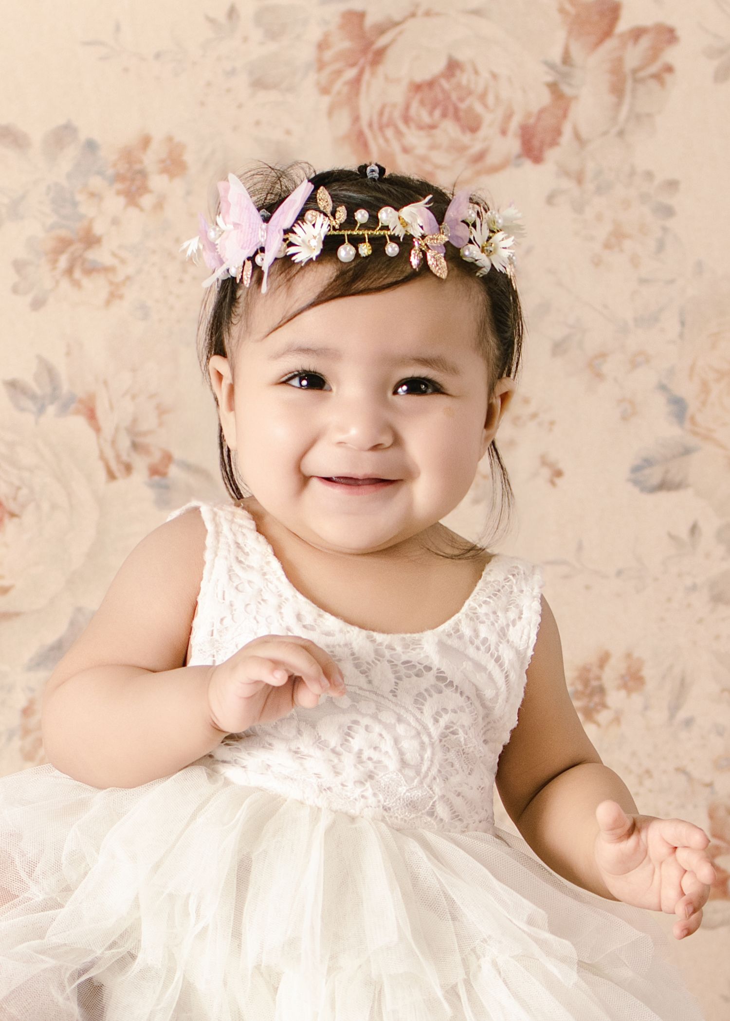 Smiling baby girl in white lace dress and floral crown poses against a soft floral backdrop.