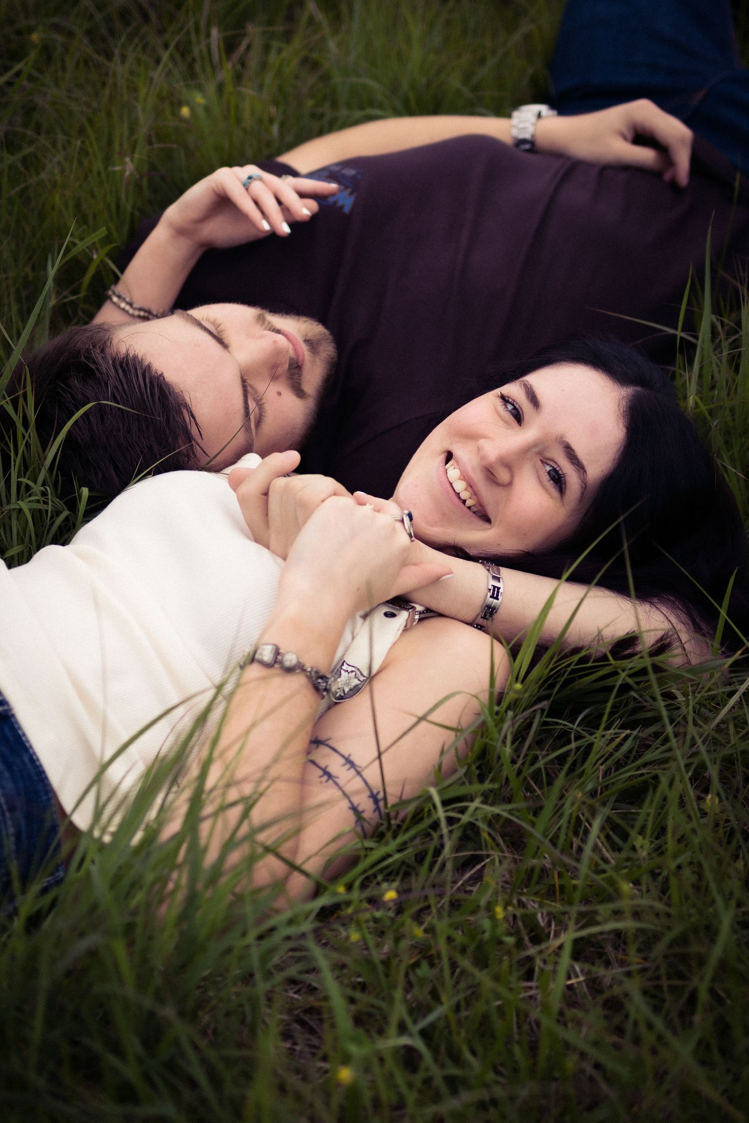 A smiling couple lying together in lush green grass, holding hands and enjoying a romantic outdoor moment.