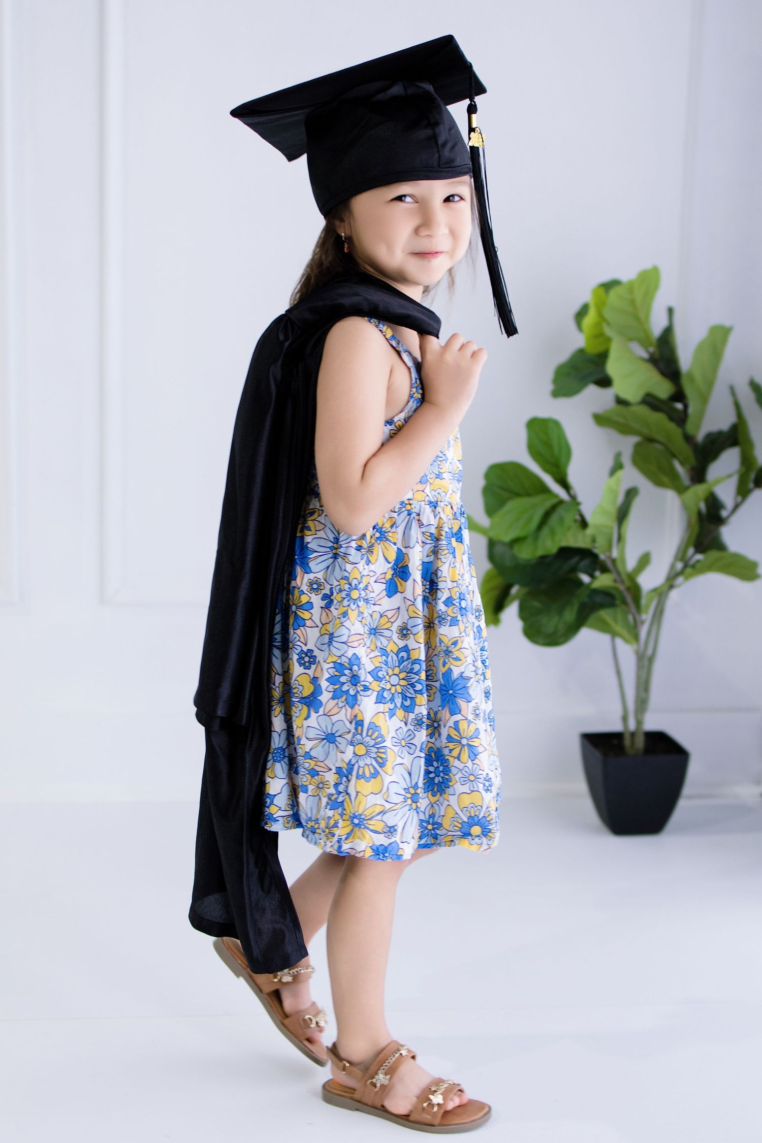 Young girl in floral dress wearing black graduation cap and gown, smiling over shoulder.