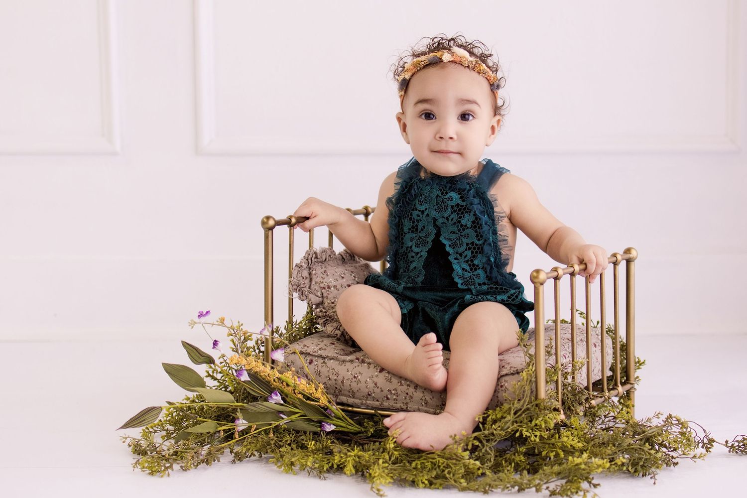 Baby girl in teal romper and floral crown sitting on gold mini bed with greenery.