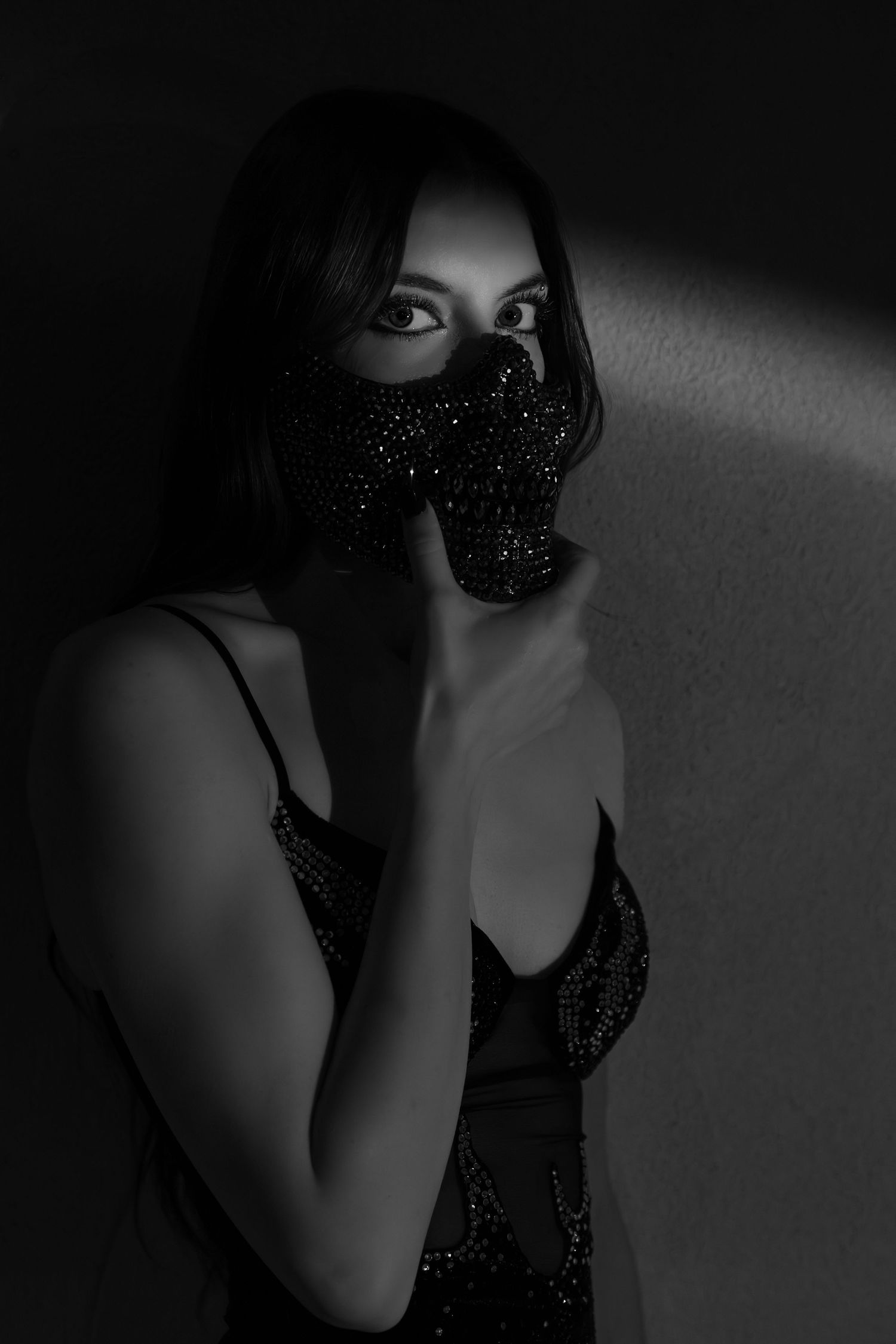 Black and white portrait of a woman wearing a sparkly face mask, leaning against a wall in dramatic lighting.