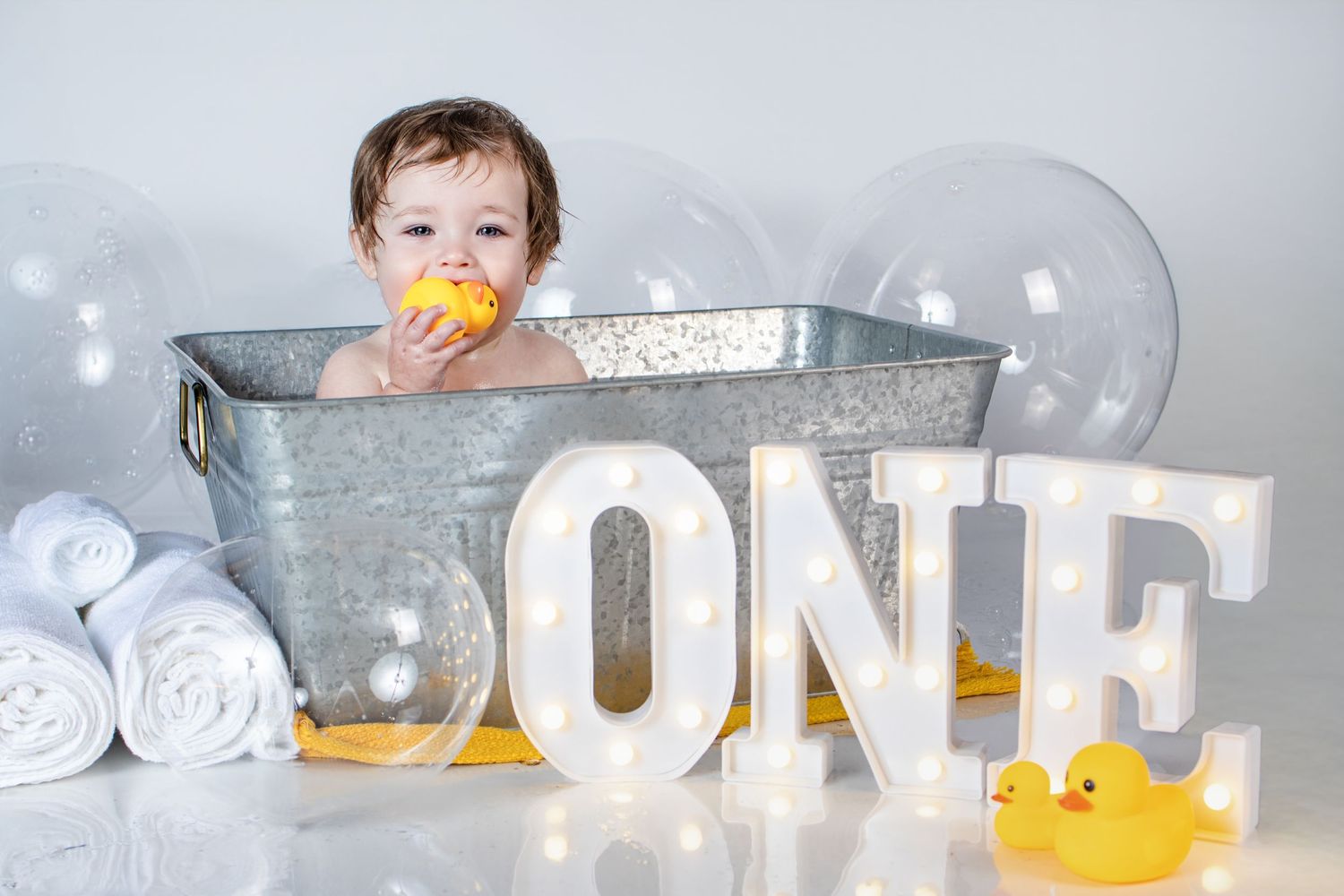 Baby sitting in a galvanized tub with rubber ducks and lit ONE letters celebrating their first birthday.