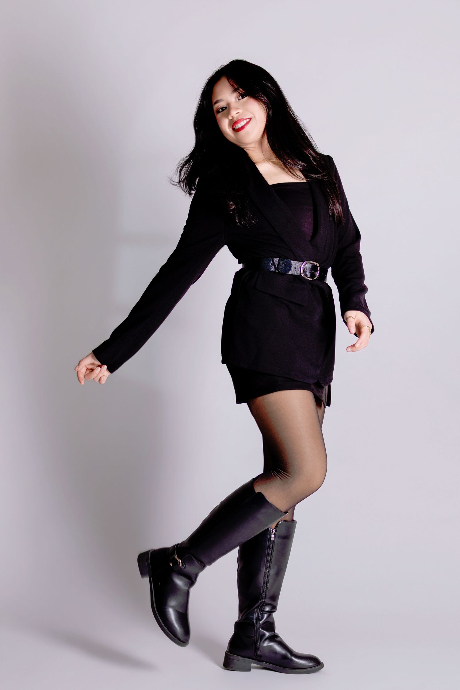 Young Asian woman in black belted mini dress and knee-high boots dancing playfully in photo studio.