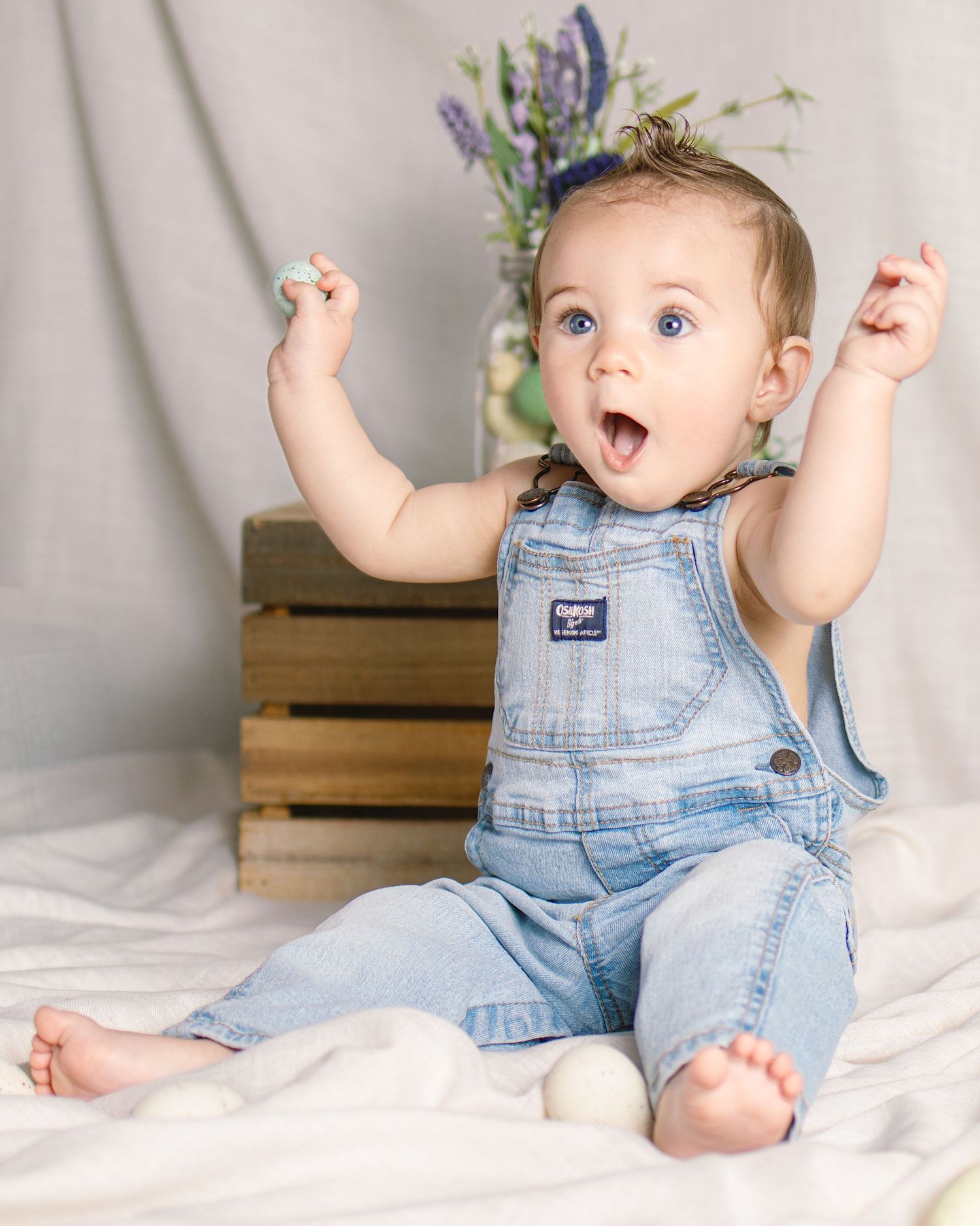 Excited baby in denim overalls sits with arms raised and mouth open wide during an Easter photo session.