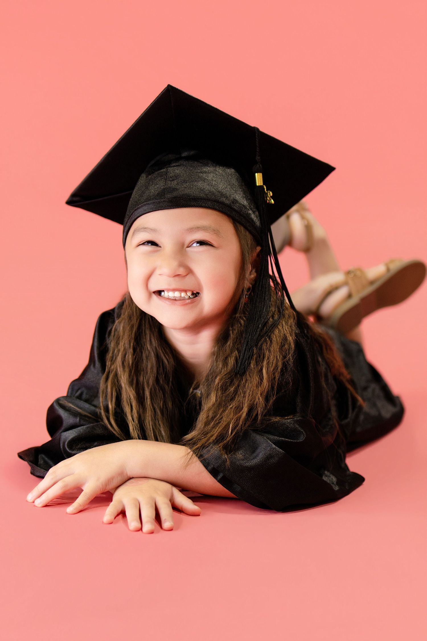Cheerful little girl in black graduation cap and gown lying down on pink background smiling.