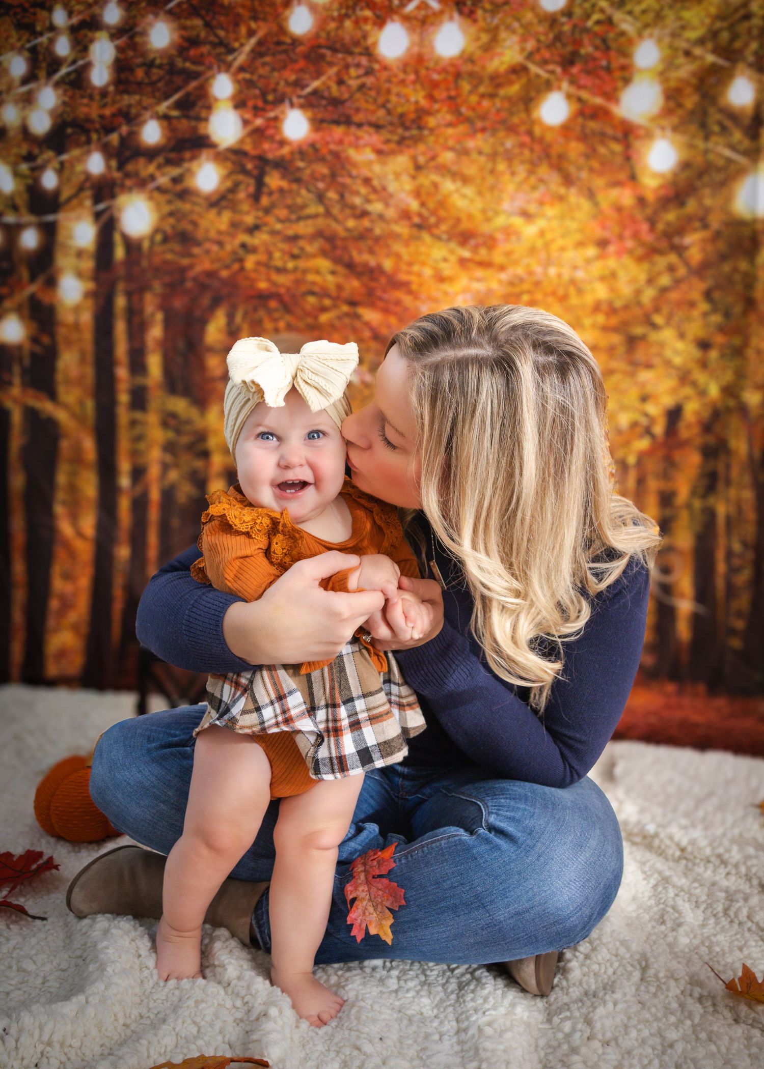 Mother kissing her smiling baby girl during a fall-themed photo session with autumn forest backdrop.