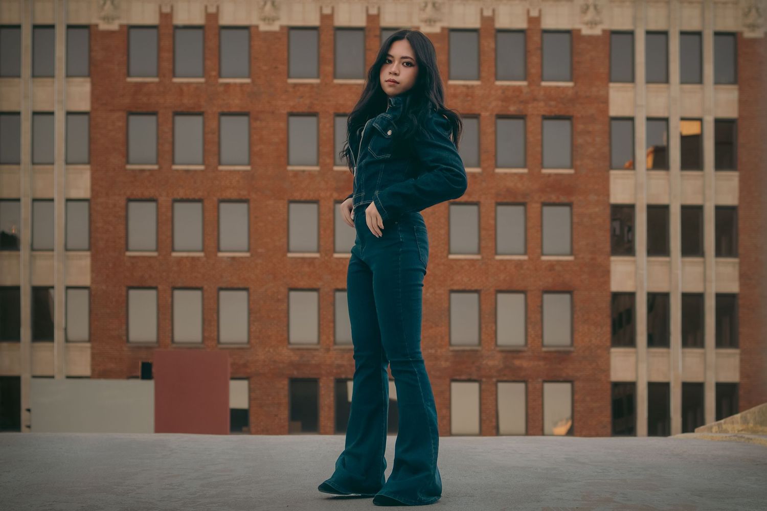 A young woman in a dark teal outfit poses confidently in front of a brick building on a rooftop.