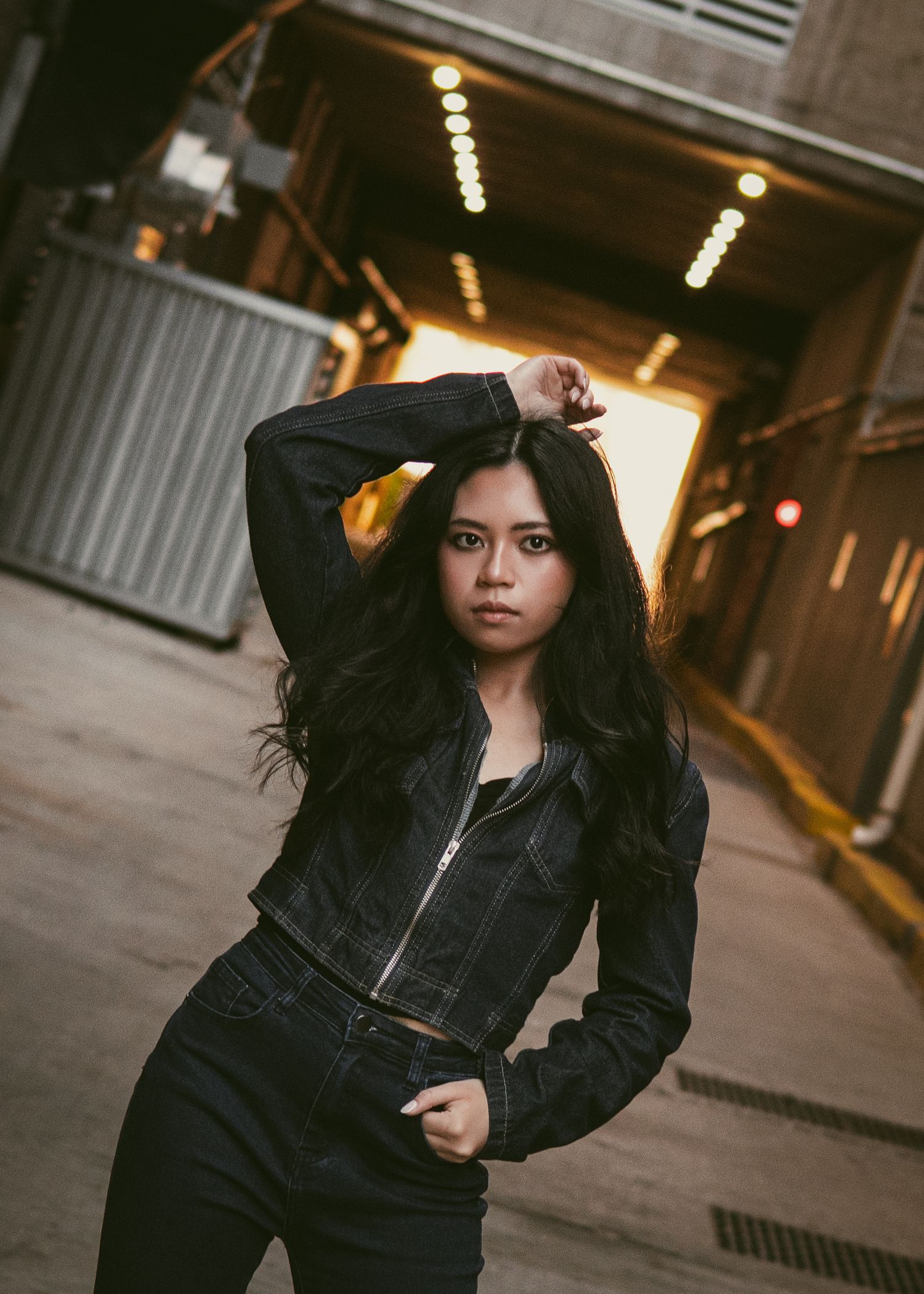 Young woman in black denim jacket posing confidently in urban tunnel with warm glowing lights.