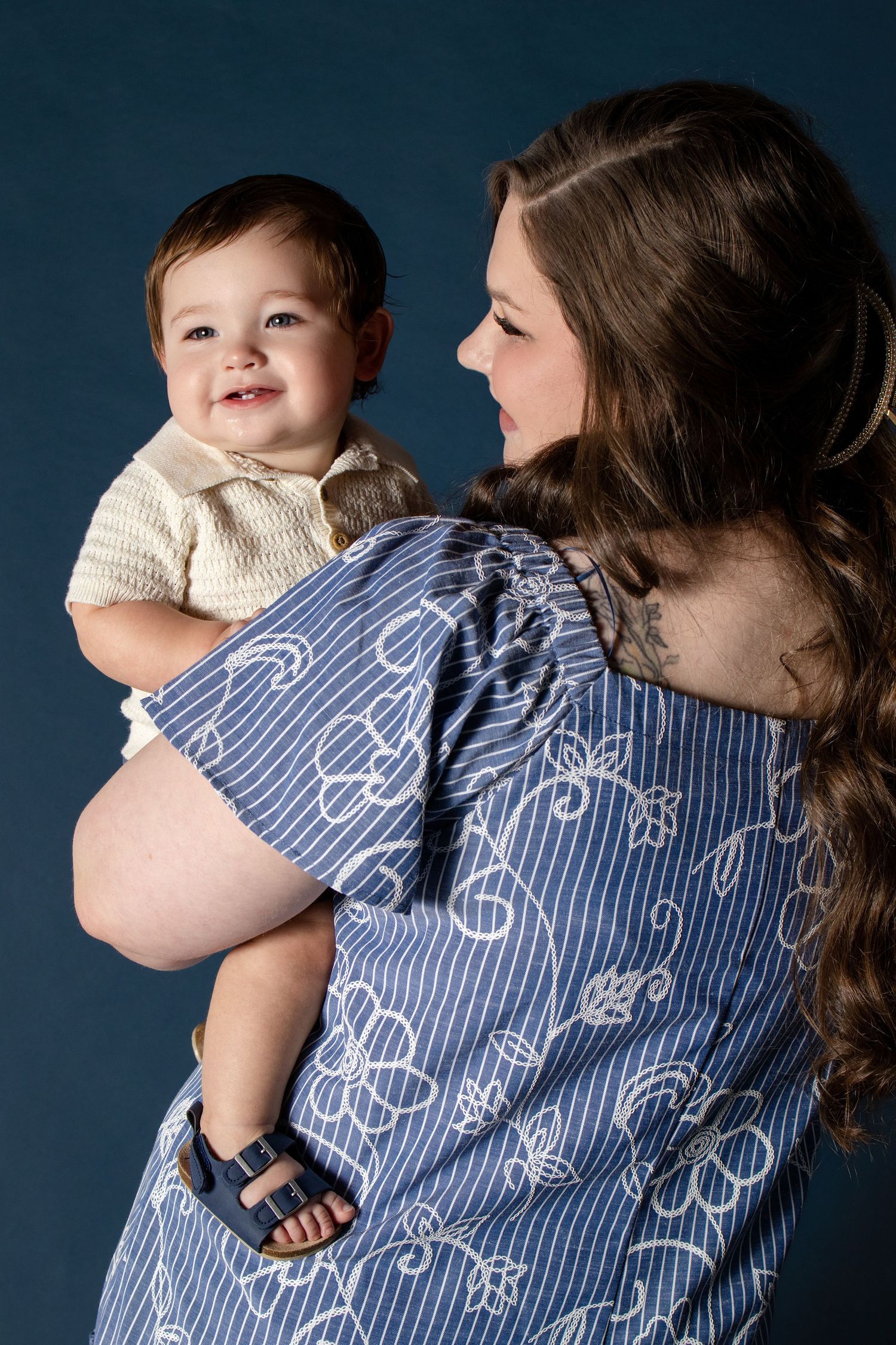 A mother in a blue patterned dress lovingly holds her smiling baby boy against a dark blue backdrop.