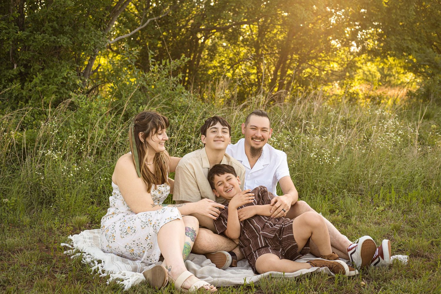 Family of four laughing together on a blanket in a sunny green meadow during a golden hour outdoor photo session.