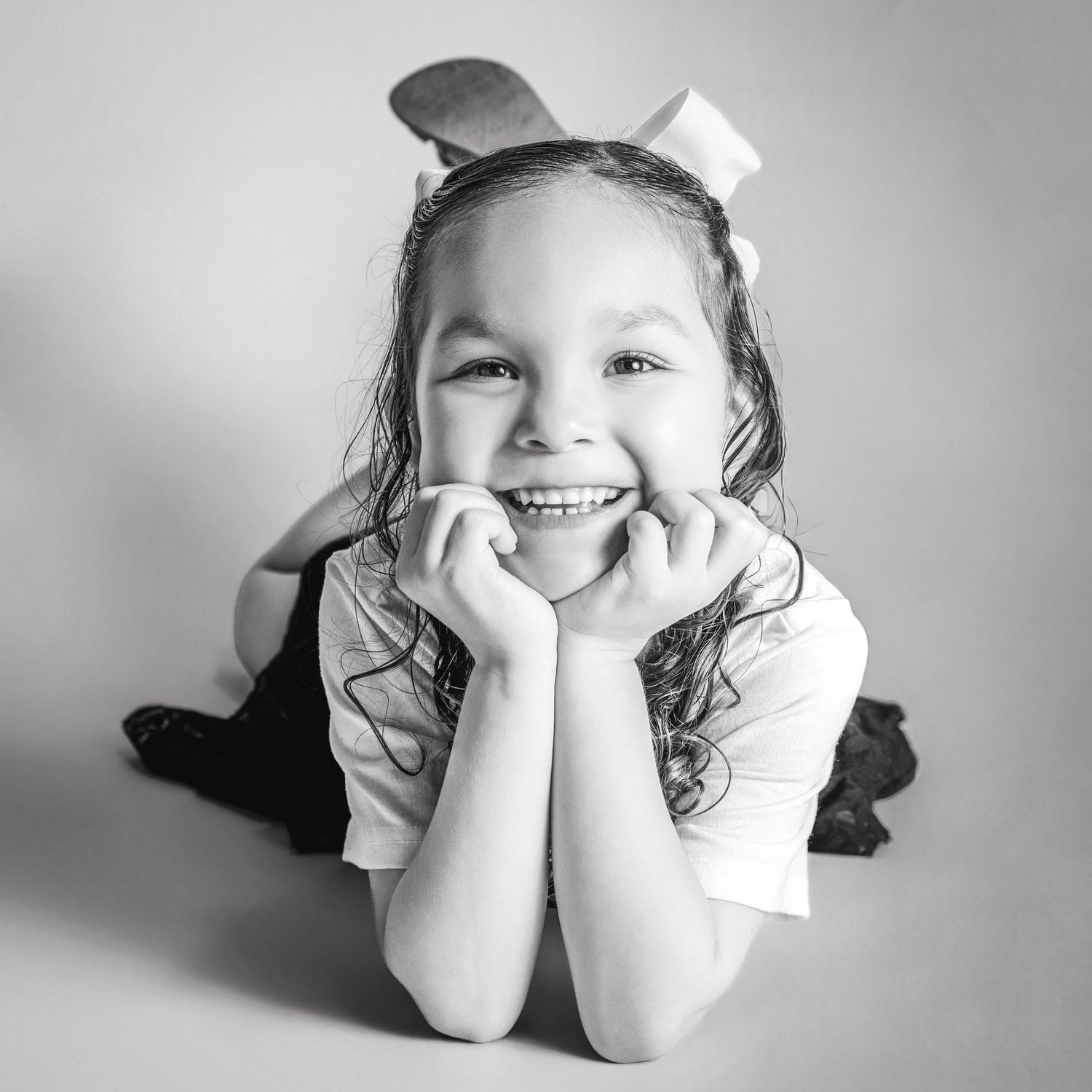 Black and white portrait of a smiling young girl lying down with hands on cheeks, wearing a bow in her hair.