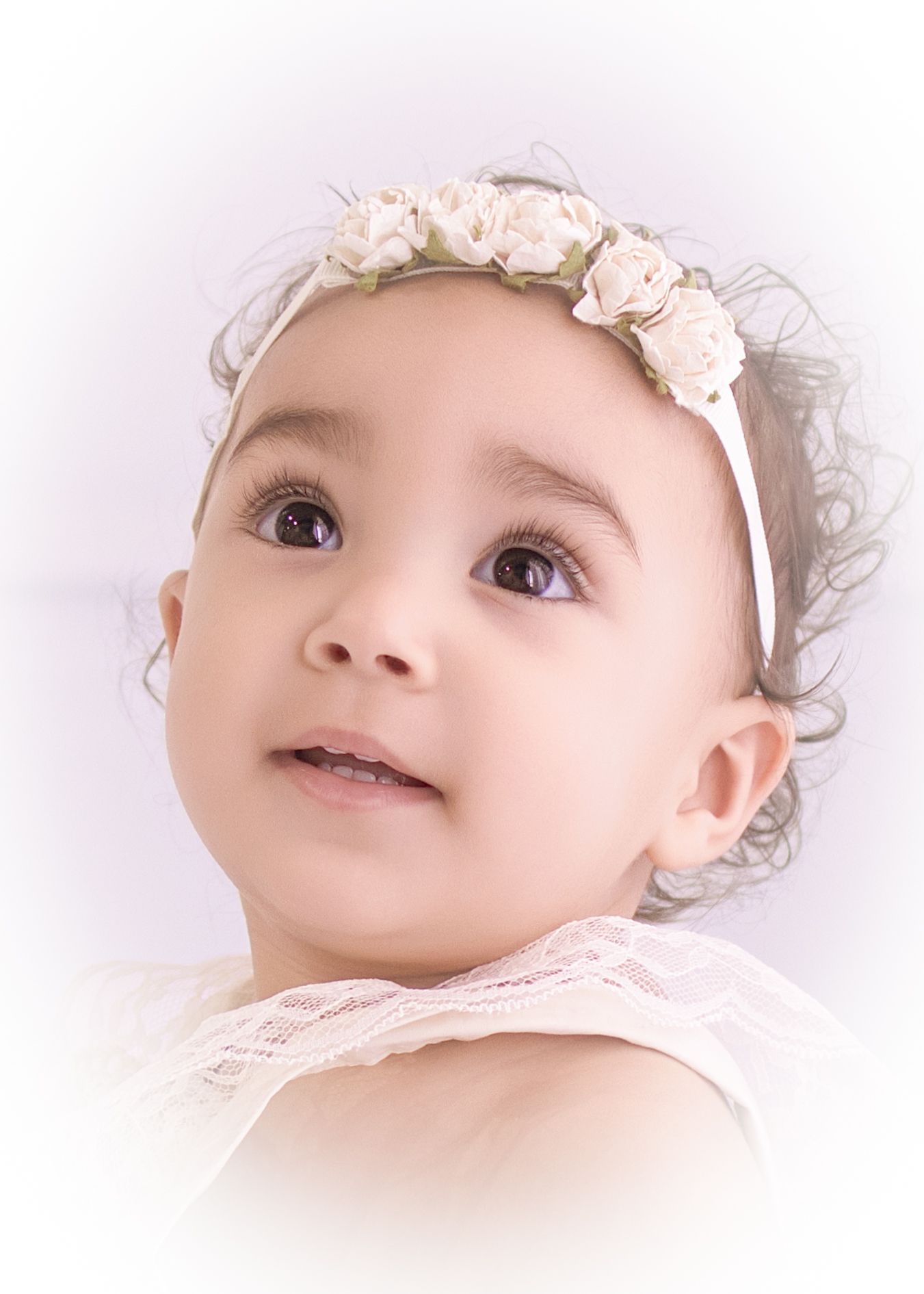 Adorable baby girl wearing a white floral headband and lace dress, looking up with big brown eyes.