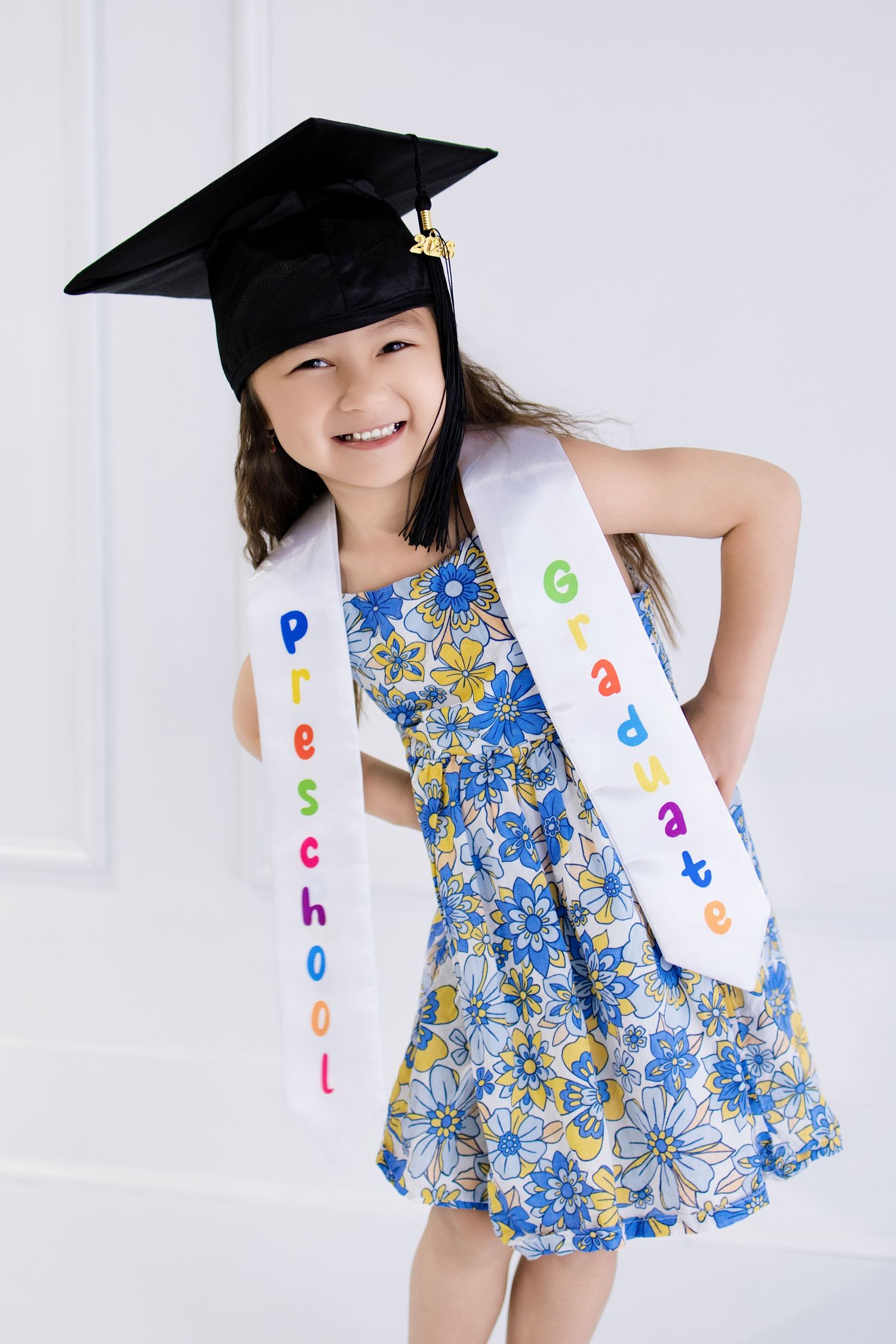 Smiling young girl wearing a graduation cap and preschool graduate sash over a floral dress, celebrating her milestone.