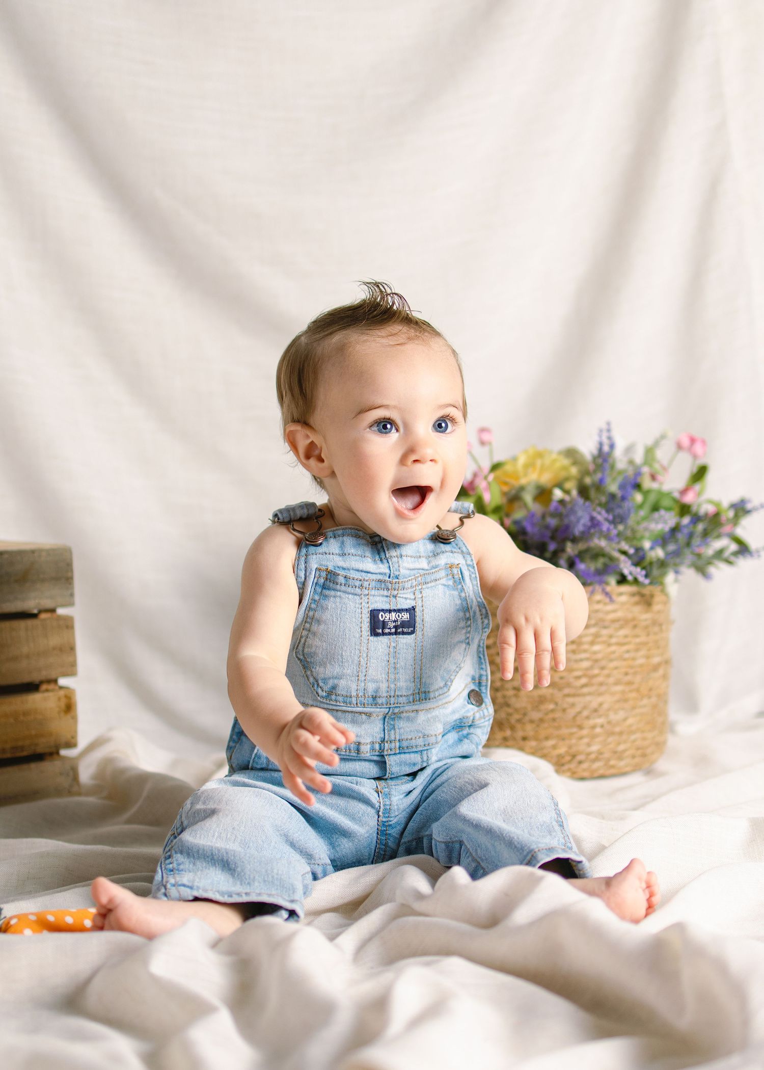 Happy baby in denim overalls sitting on white fabric with flowers and wooden crates in background.