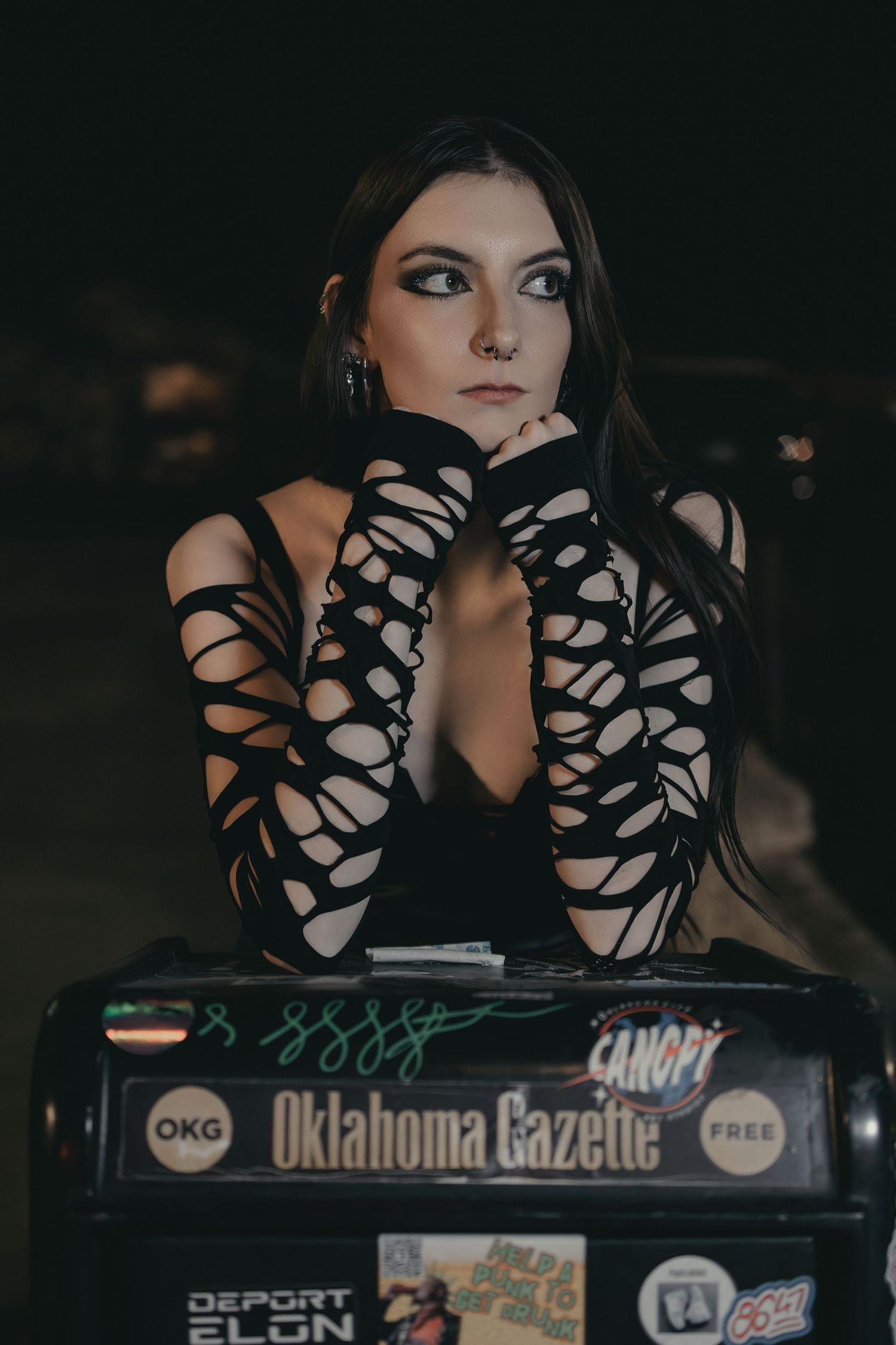 Woman in black cutout gloves leaning on Oklahoma Gazette newspaper box at night.