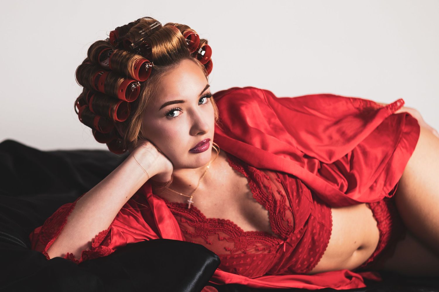 Young woman in red lace lingerie and satin robe with hair curlers poses on a dark sofa.