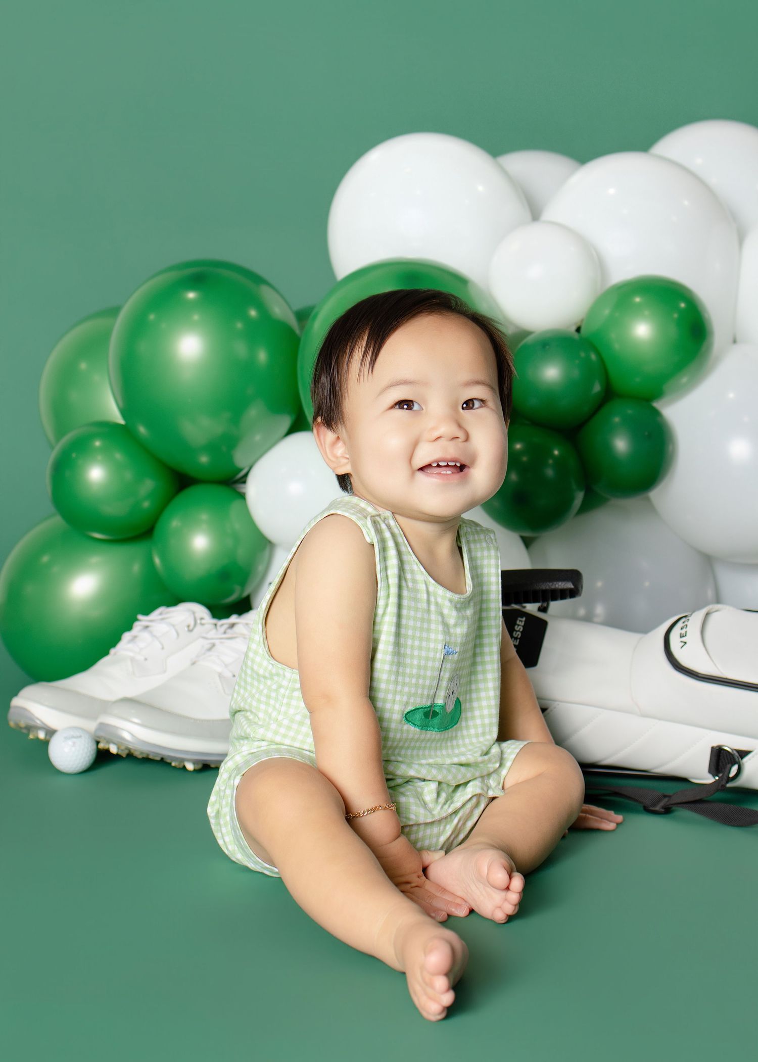Smiling baby in green gingham outfit sits before green and white balloon garland on green backdrop.