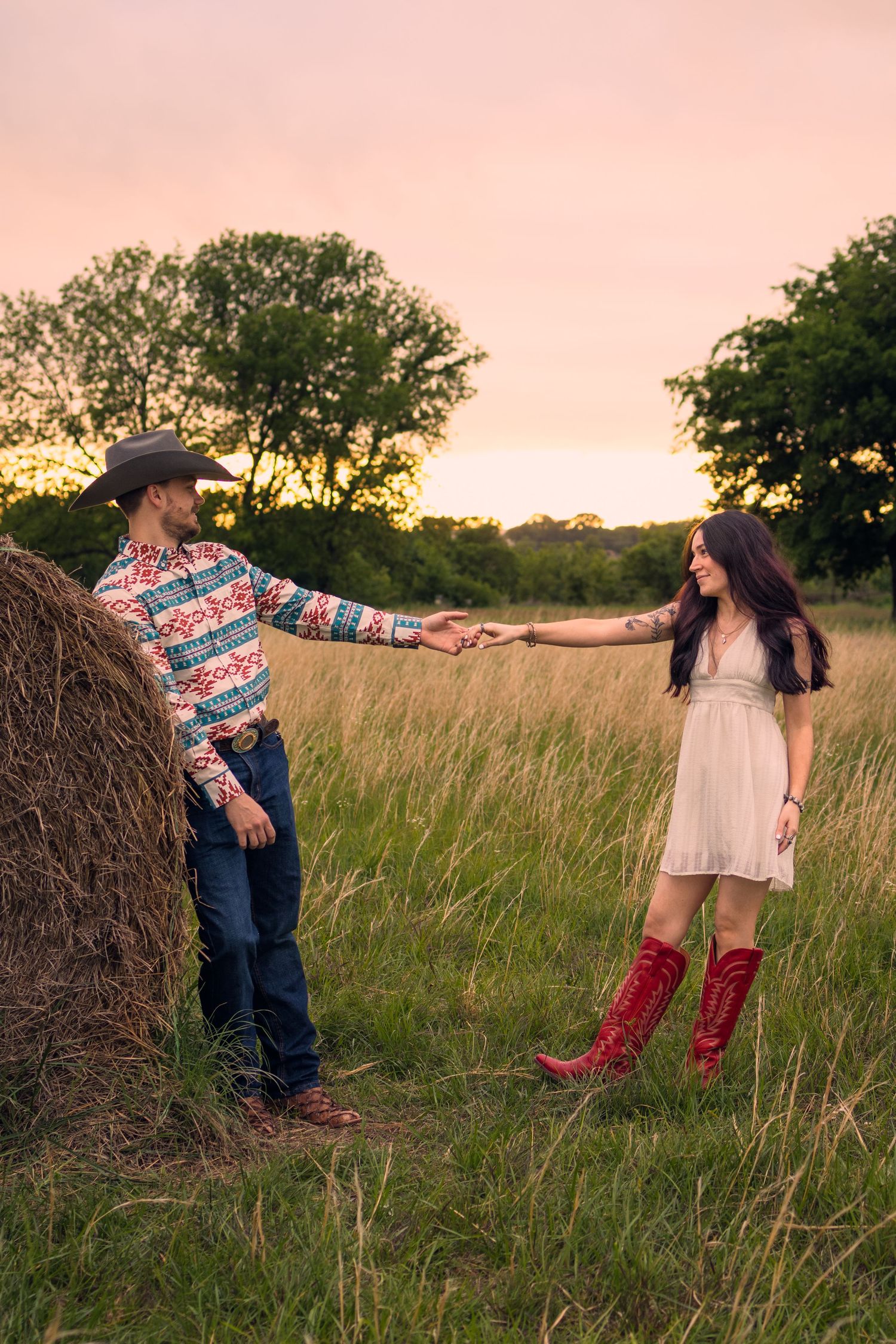 Couple in western attire holding hands in a golden field at sunset near a hay bale.
