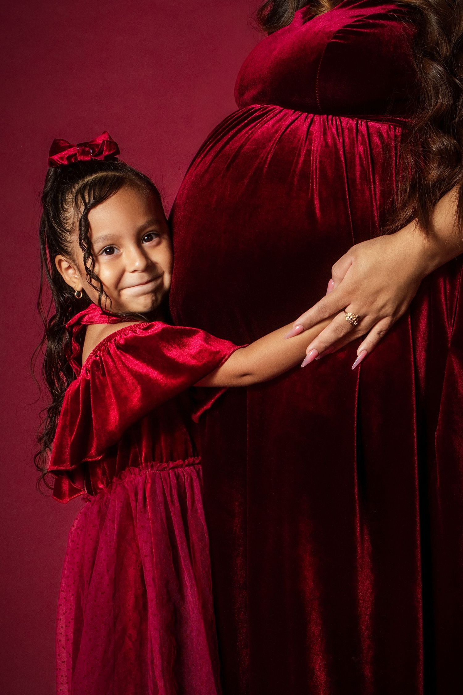 A young girl in a red velvet dress hugs her pregnant mother's belly during a maternity photo session.