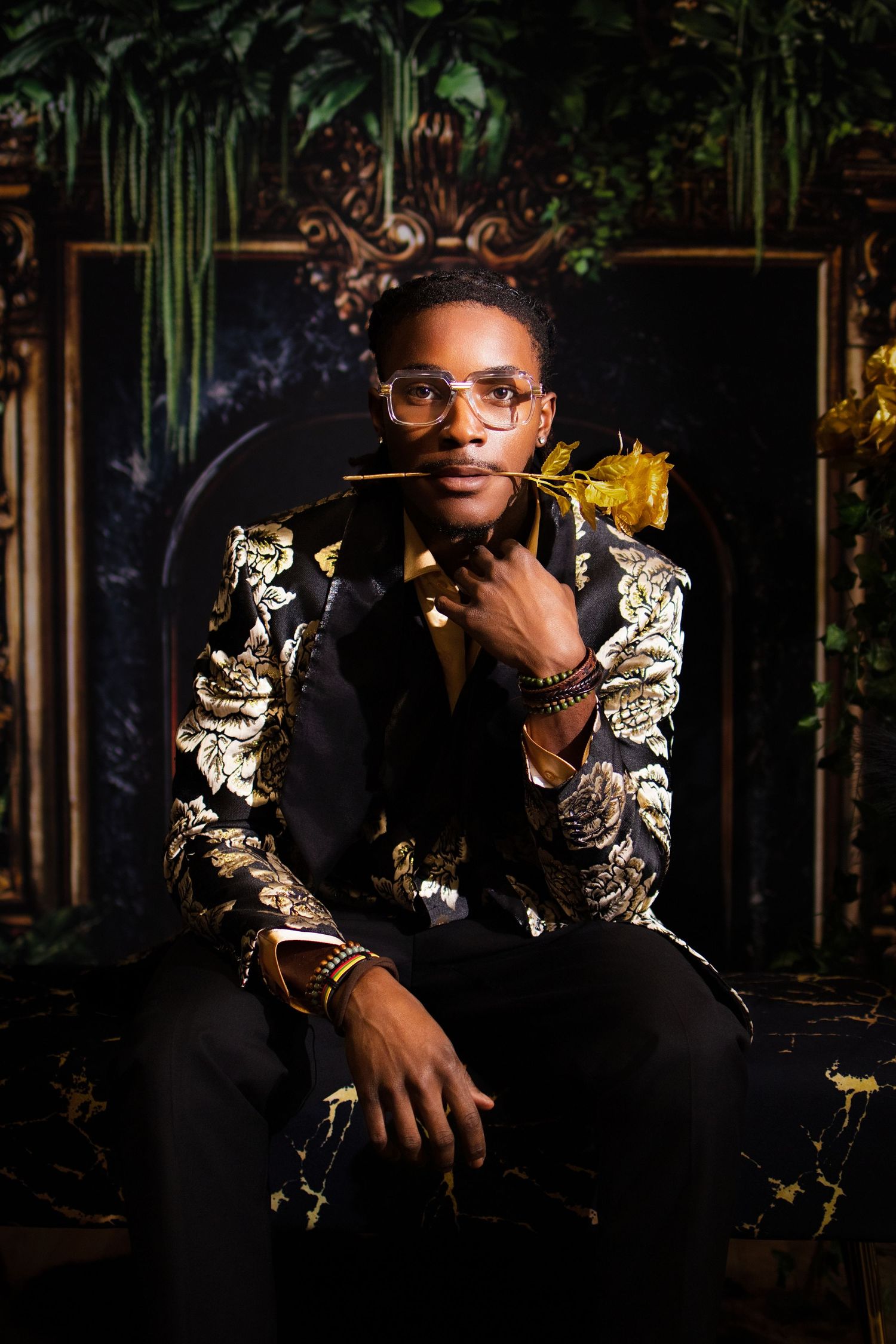 A stylish man in a gold floral blazer holds a yellow rose, wearing glasses, seated before an ornate dark backdrop.