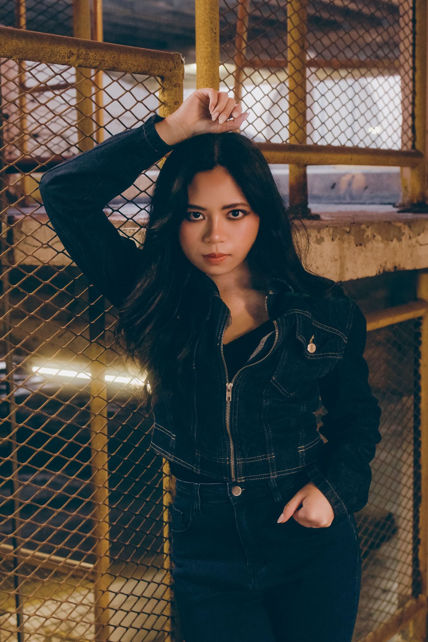 Young woman in black denim jacket poses against chain-link fence in urban setting.