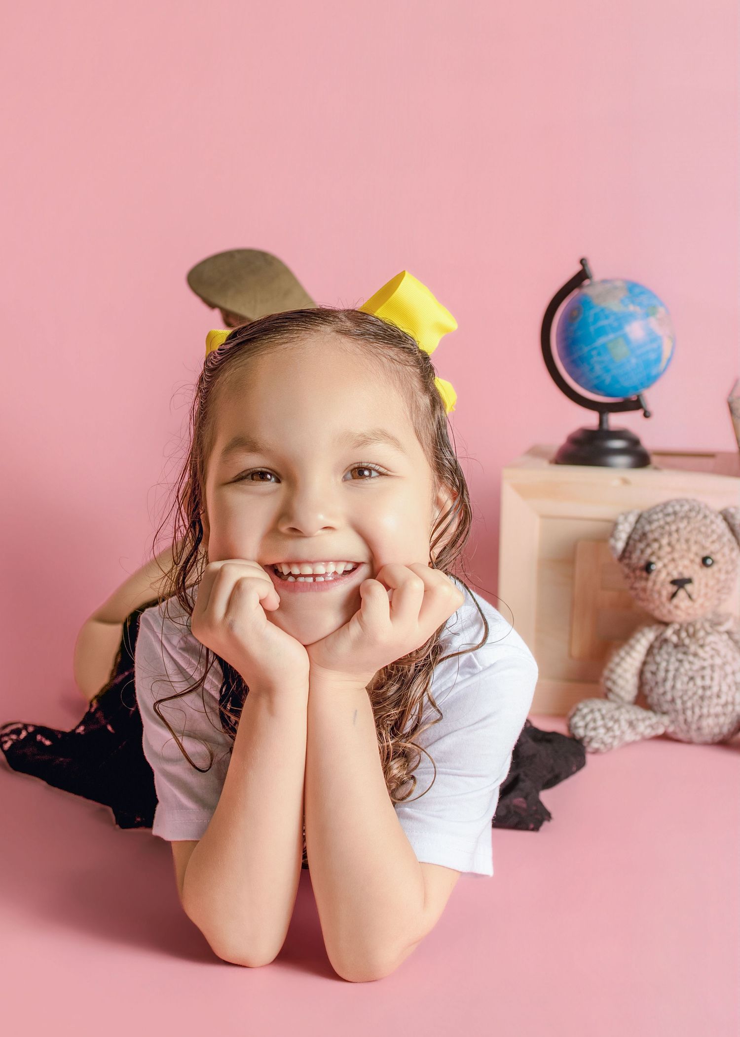 Smiling young girl lying on pink floor with hands on cheeks, yellow hair bows, globe and teddy bear in background.
