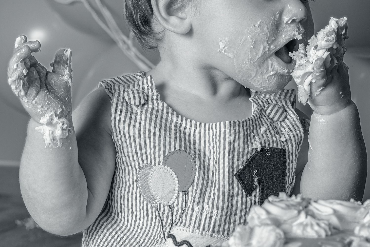 Black and white photo of a toddler in striped dress enjoying a cake smash, with frosting on hands and face.