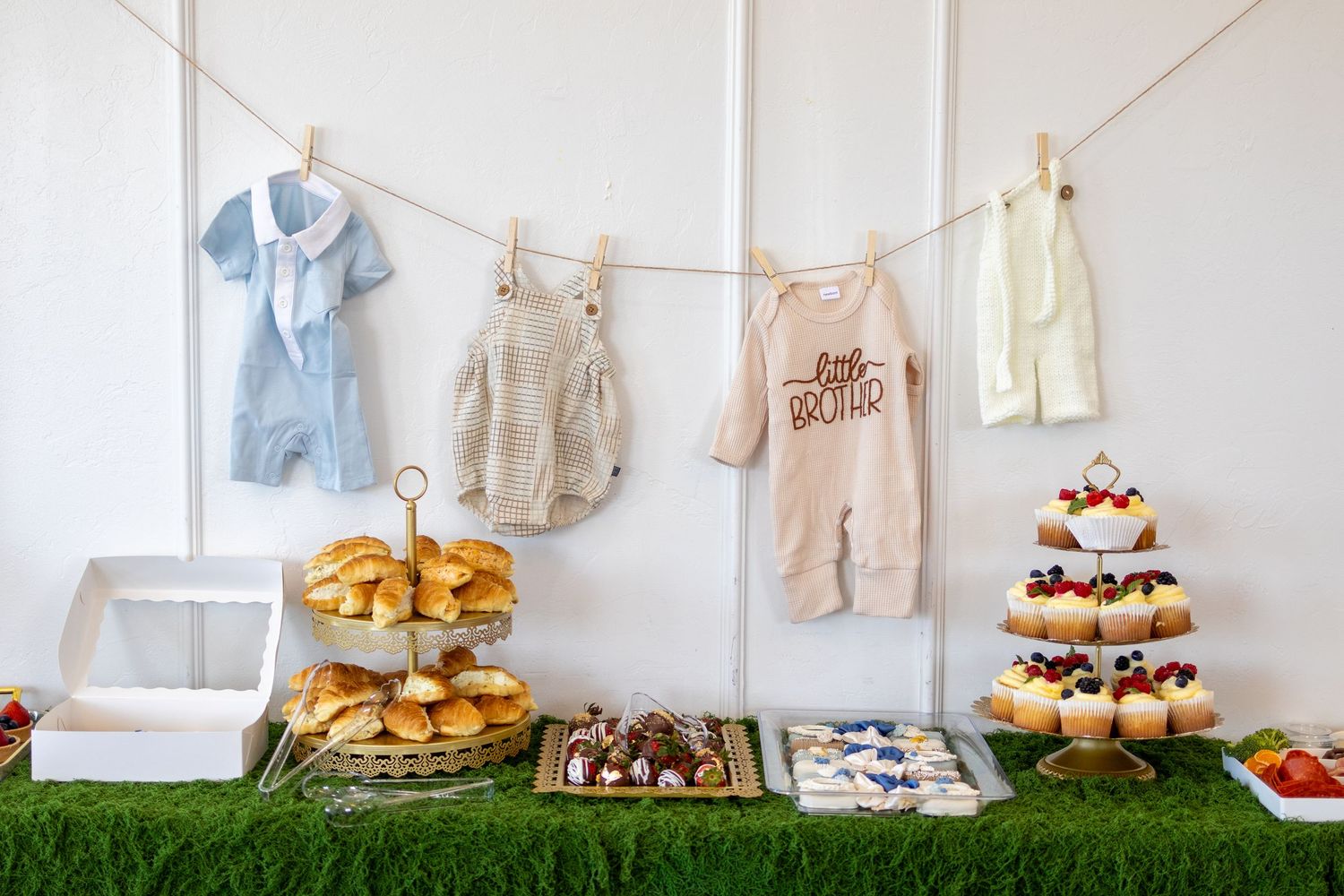 Baby shower dessert table with cupcakes, pastries, and baby clothes hanging on a clothesline above.