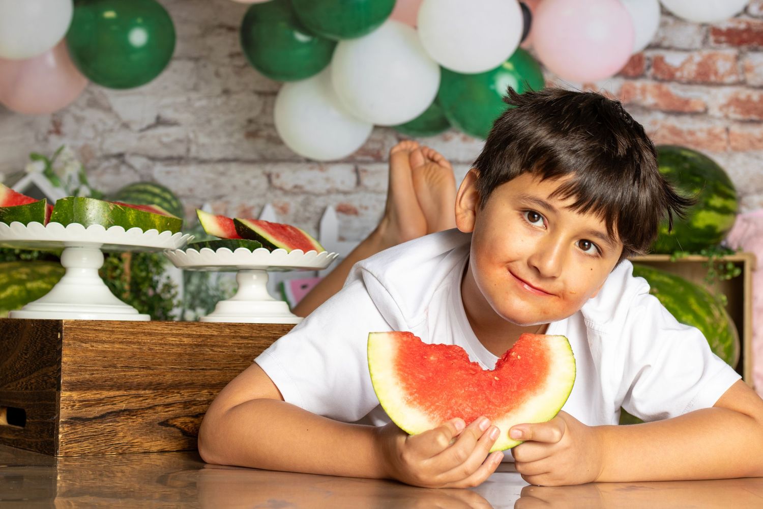 A smiling boy holds a heart-shaped watermelon slice at a festive table with balloons and fruit decorations.