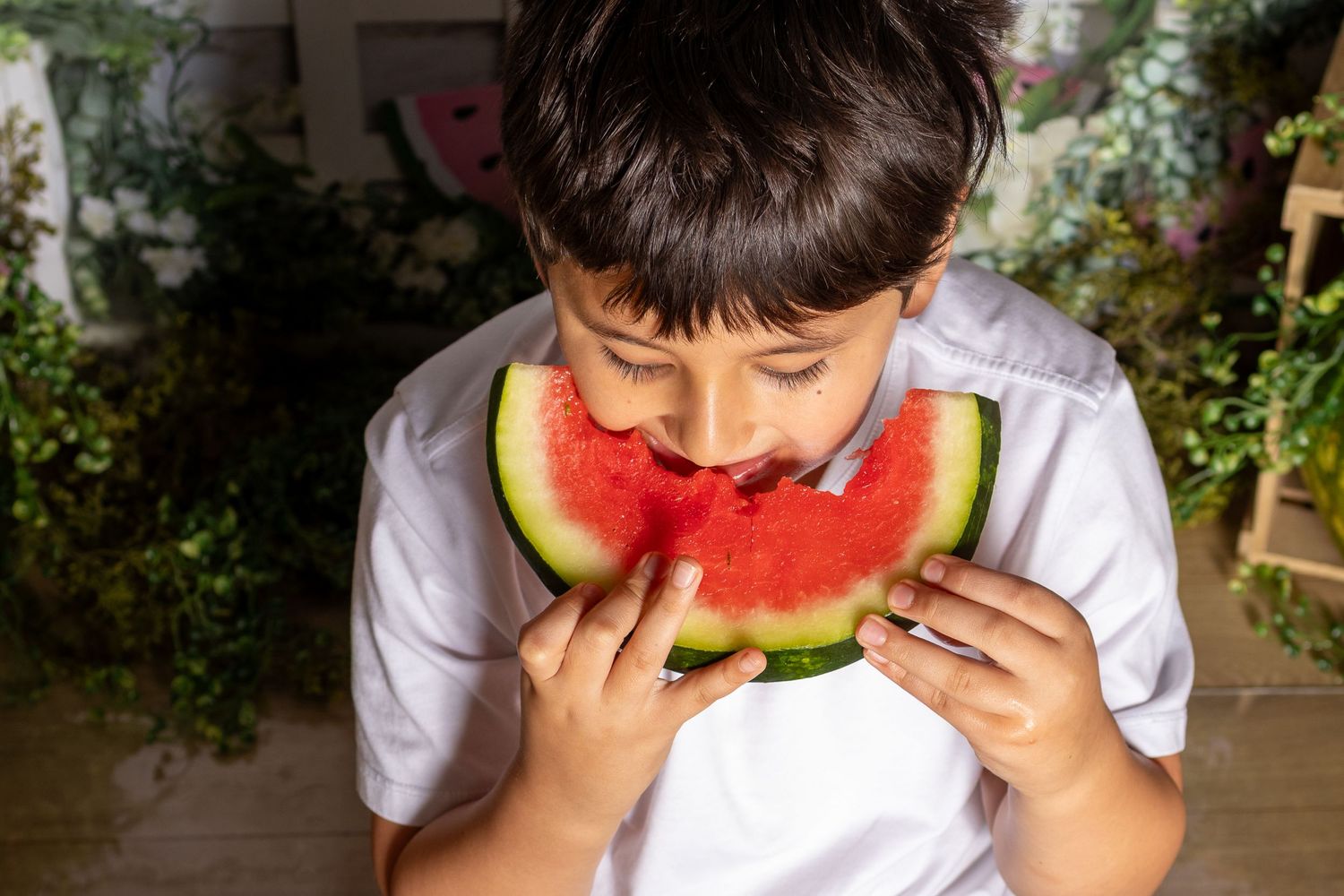 Young boy in white shirt eagerly eating a fresh slice of red watermelon outdoors.