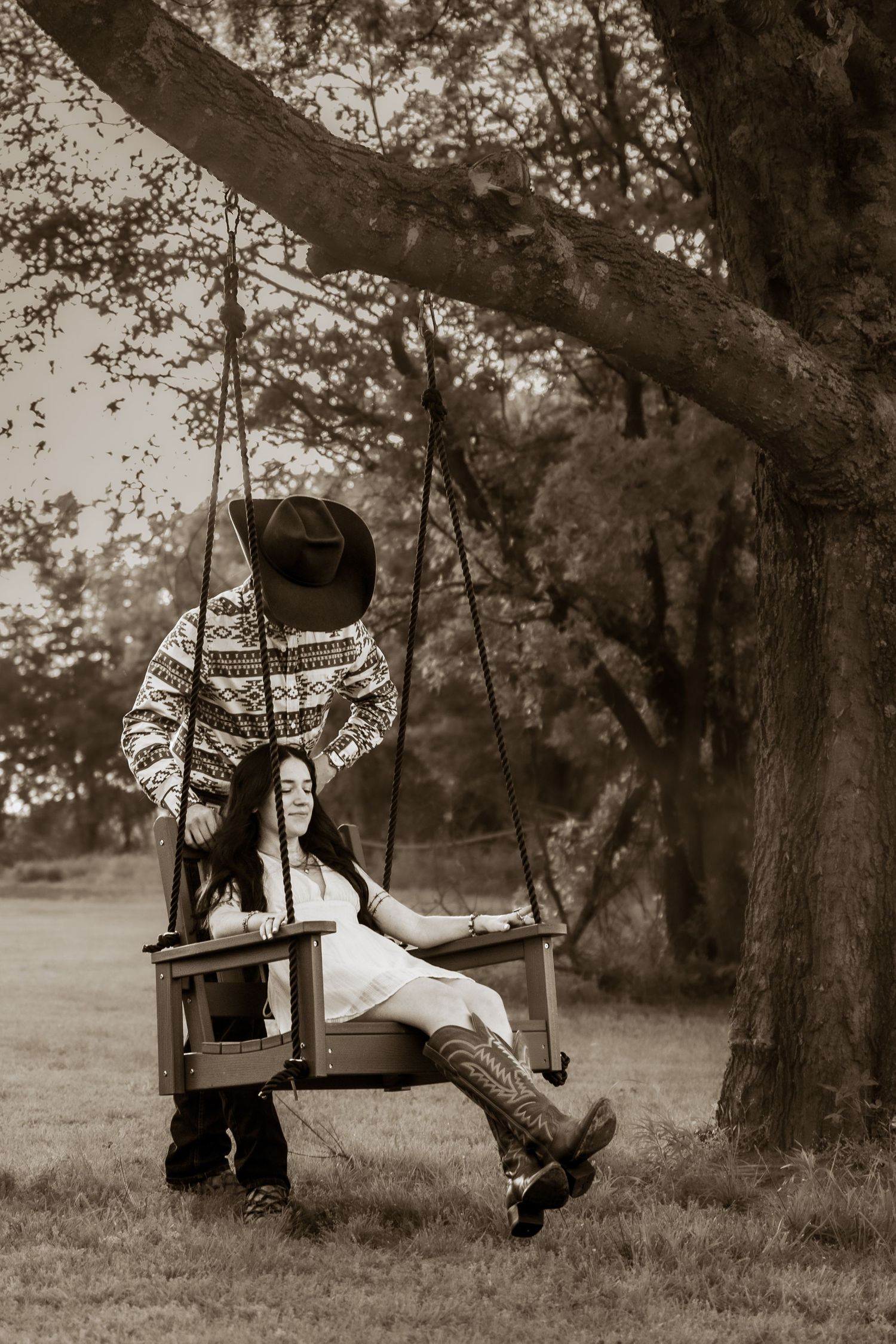 Sepia-toned photo of a couple, woman in boots sitting on a tree swing while man in cowboy hat stands behind her.