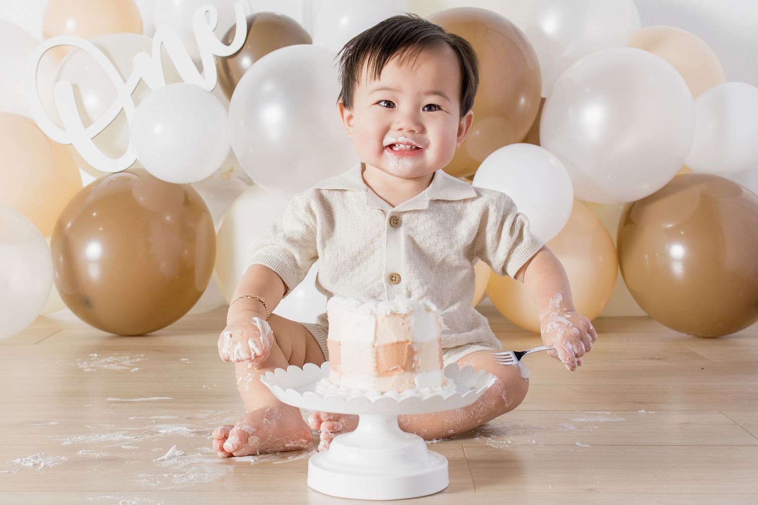 Smiling baby boy in beige outfit celebrating first birthday with cake smash, surrounded by white and gold balloons.