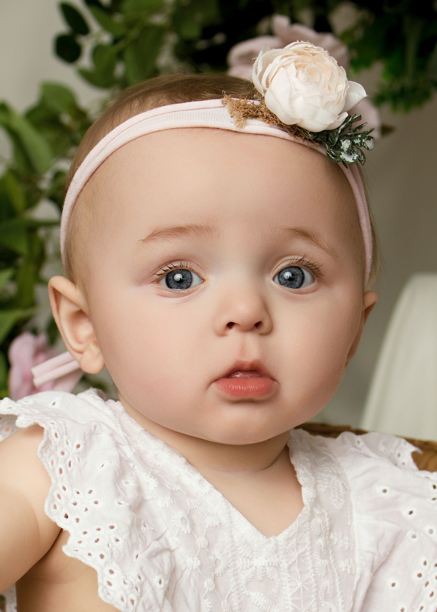 Adorable baby girl with blue eyes wearing a white eyelet dress and pink floral headband, posed in front of soft floral backdrop.