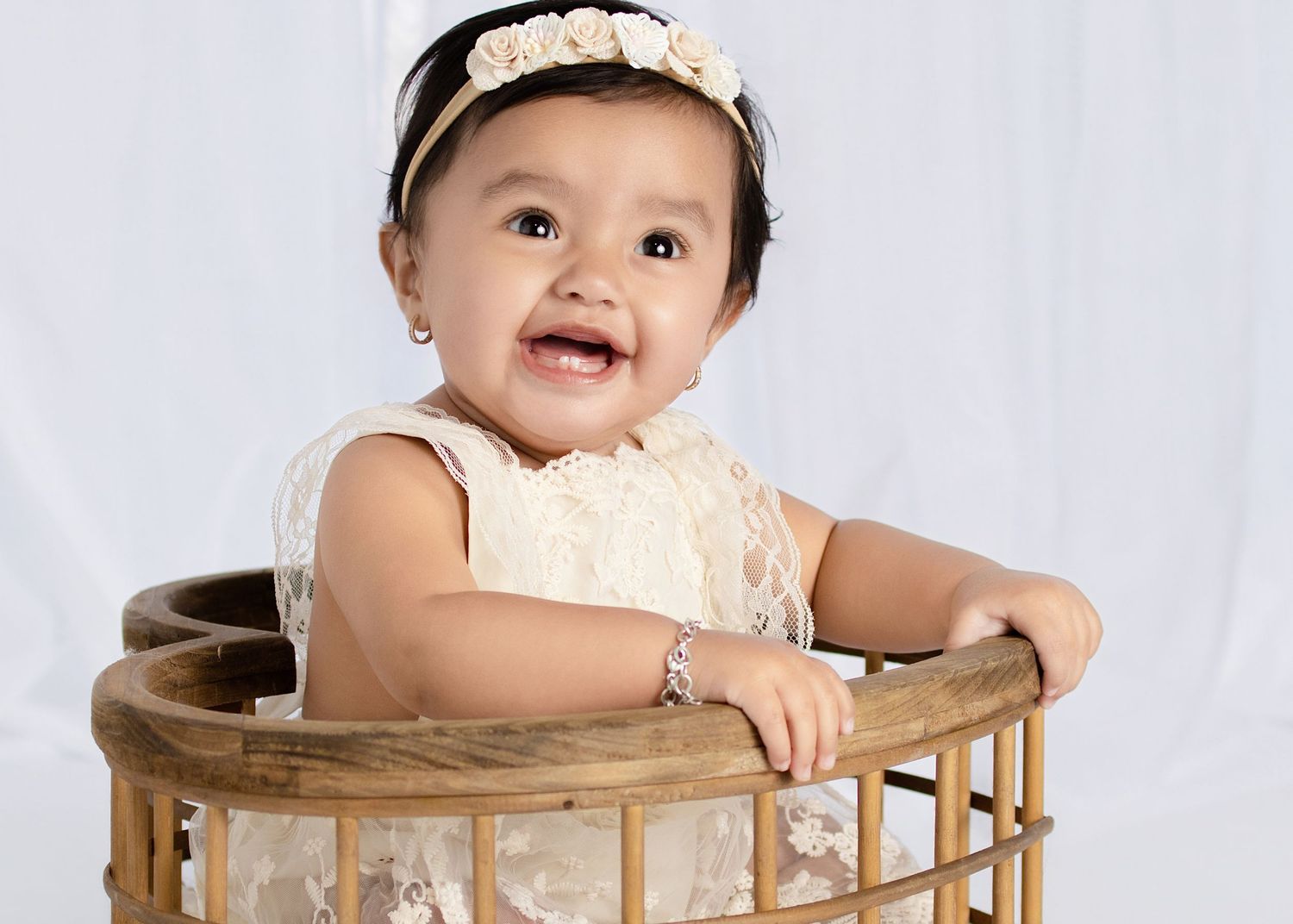 Smiling baby girl in white lace dress and floral headband sitting in a wicker basket for a portrait photo.