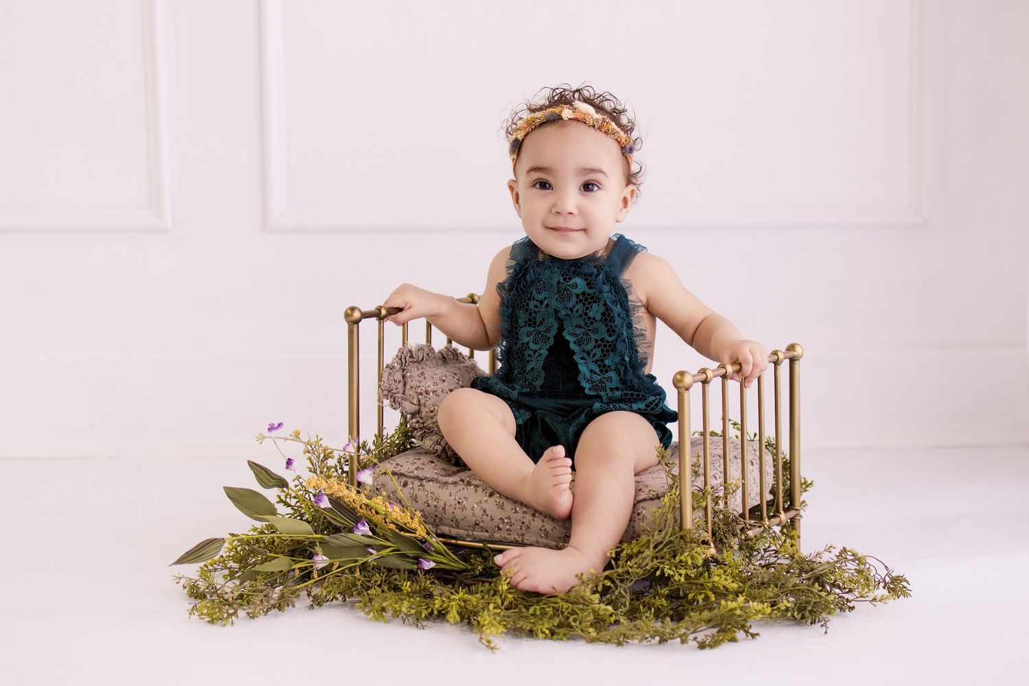 Baby girl in teal romper and floral crown sits in gold mini bed surrounded by greenery and flowers.