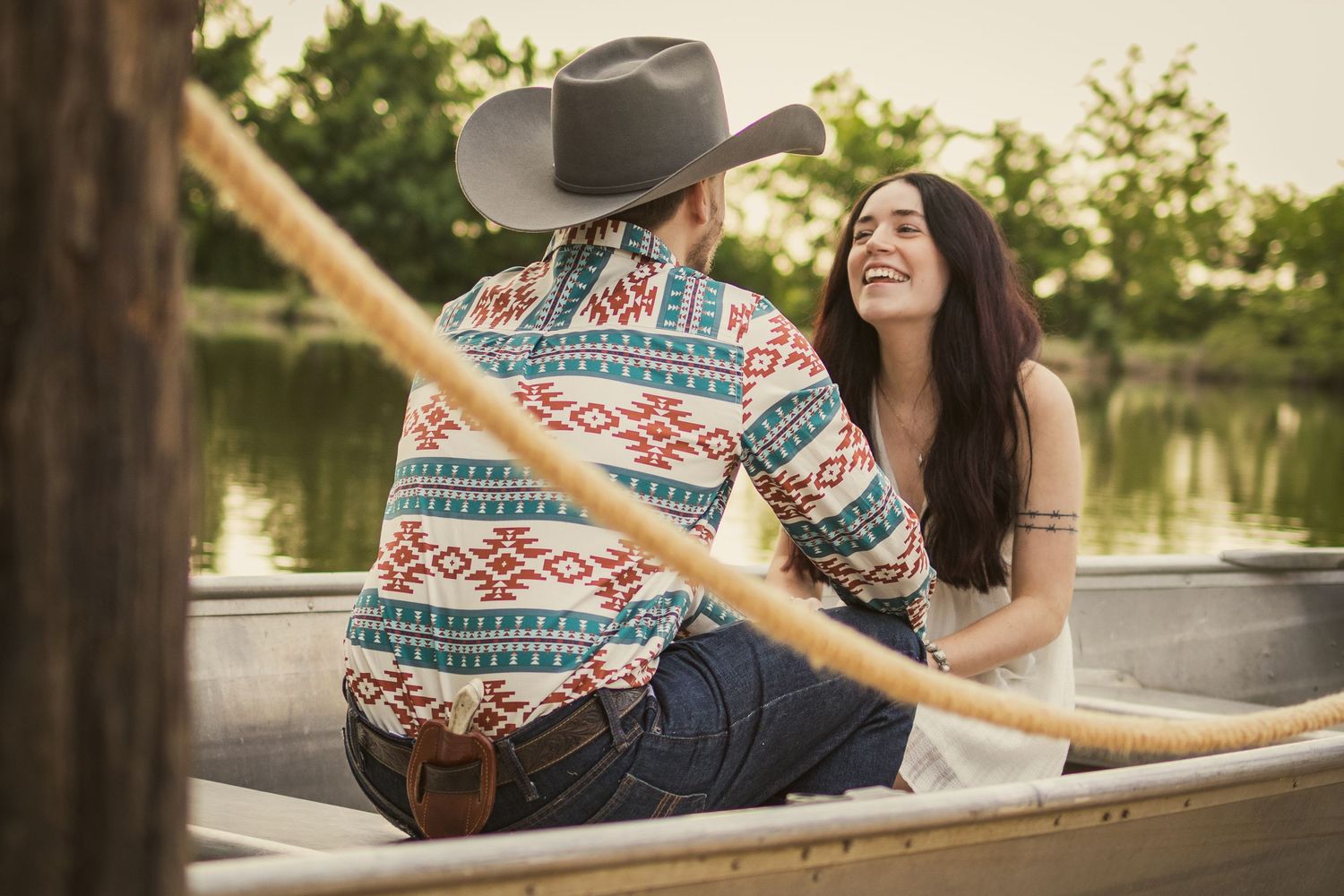 Couple sitting in a canoe on a calm lake, man in cowboy hat and patterned shirt, woman in white smiling.