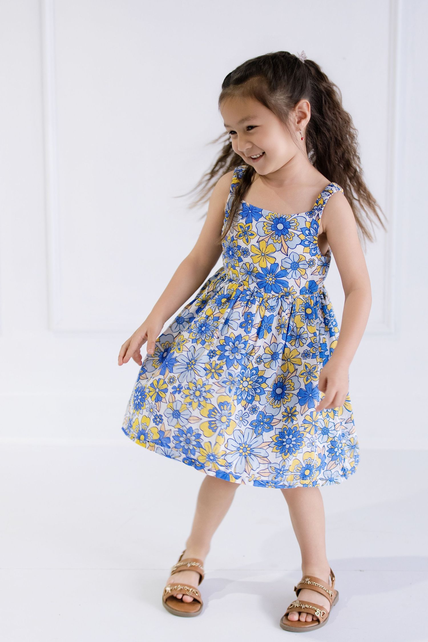 Young girl in blue floral sundress and sandals smiling and twirling in a bright white studio setting.