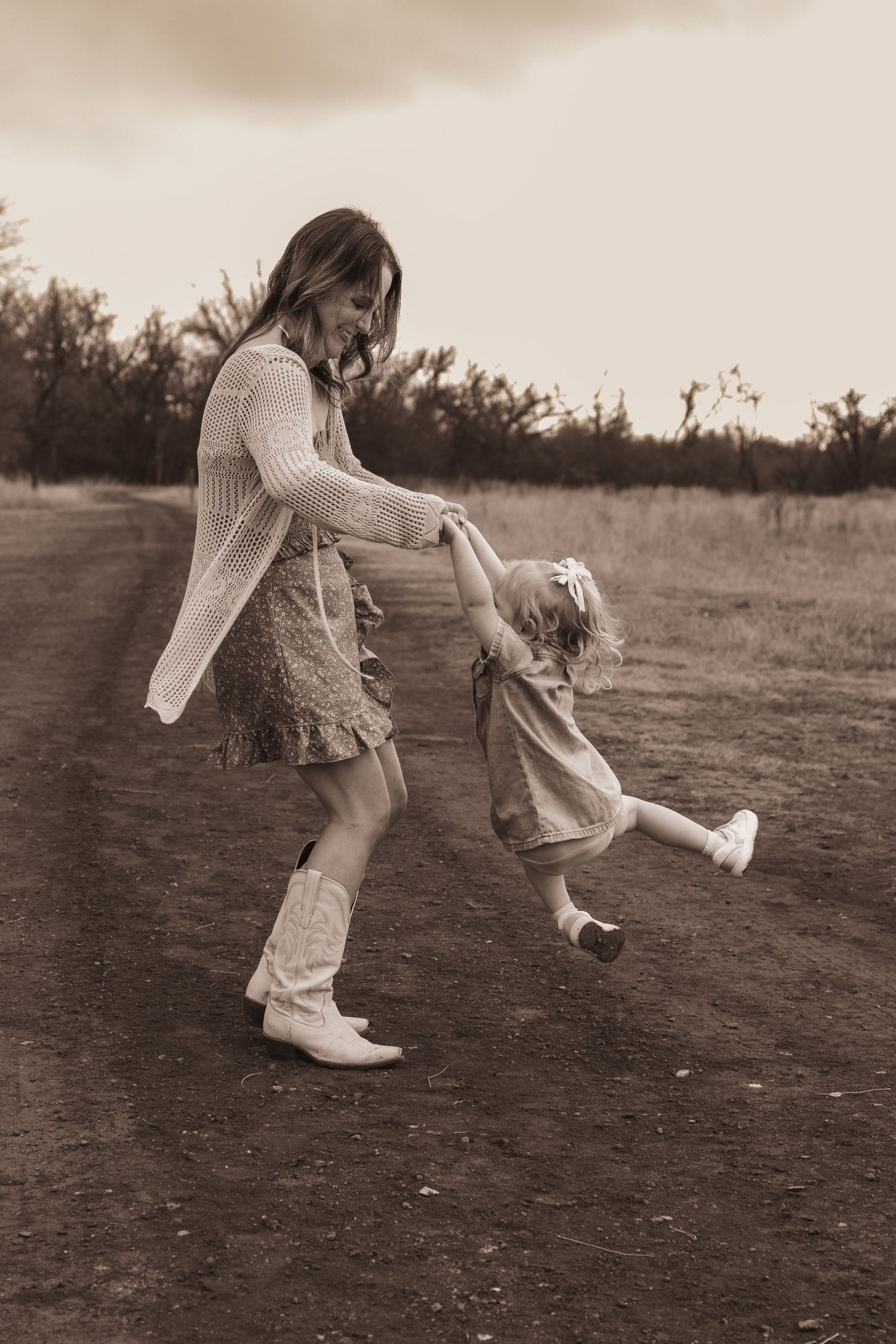 Mother and daughter dancing on a rural dirt path in sepia-toned photo, wearing dresses and cowboy boots.