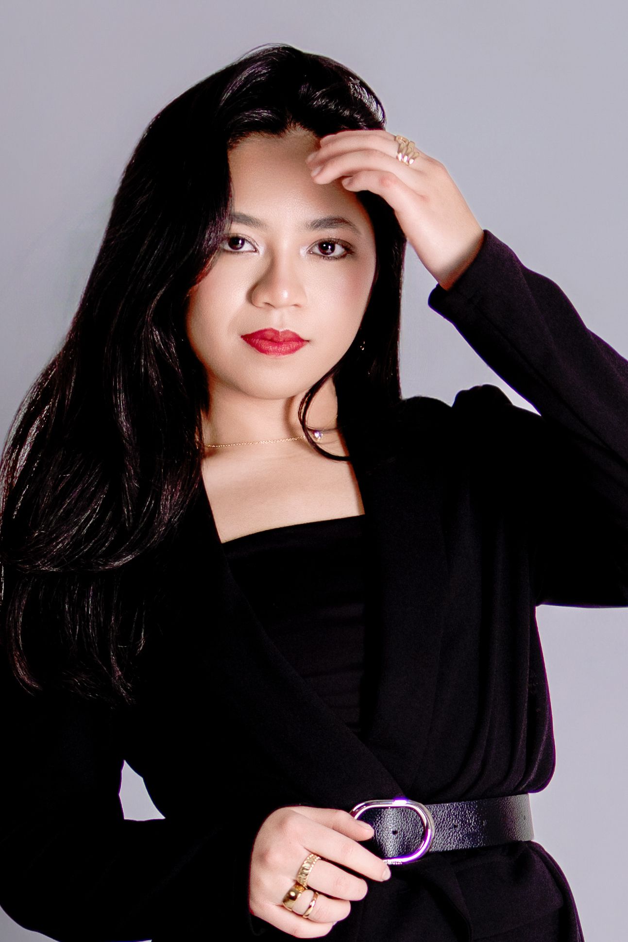 Young Asian woman with long black hair, red lips, wearing a black outfit with belt, posing with hand raised to forehead.