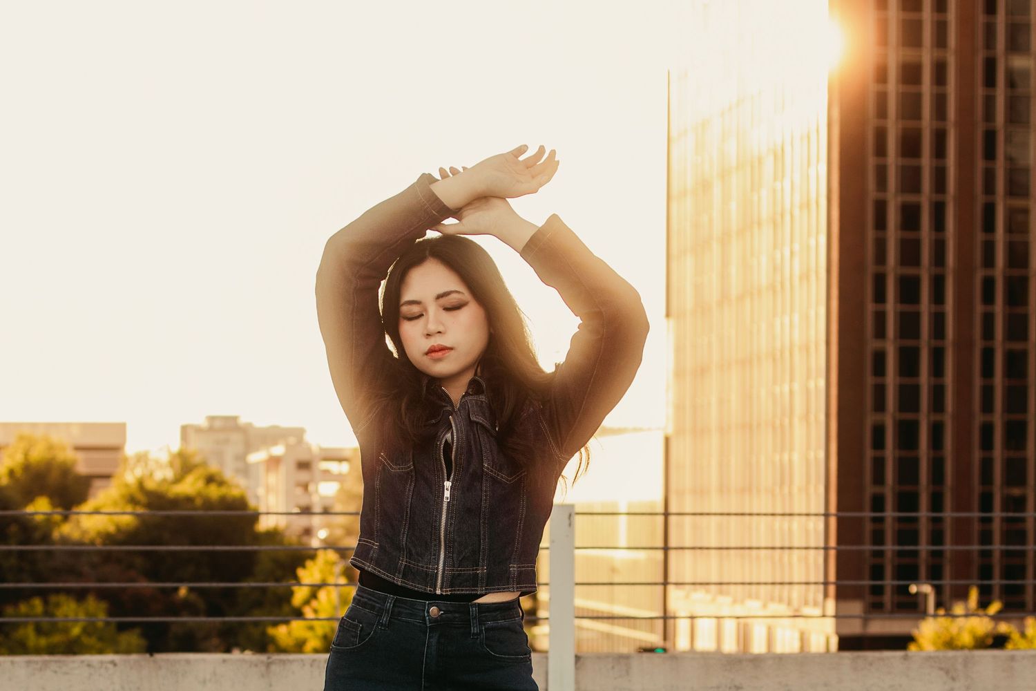 Young Asian woman poses with arms raised against golden sunset sky on urban rooftop with city buildings behind her.