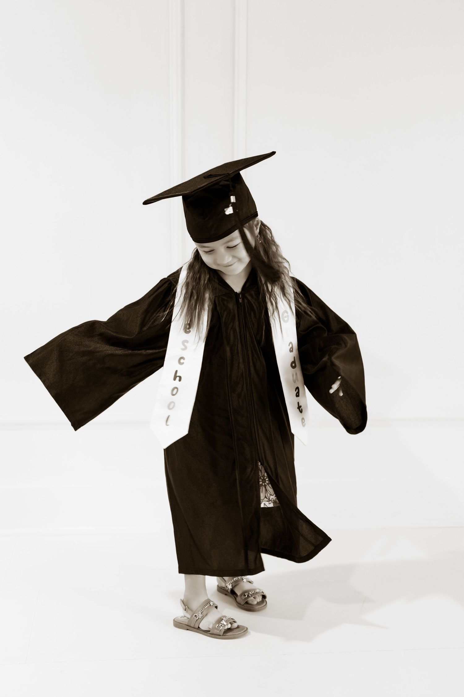 A young girl twirls in a black graduation cap and gown in a bright white studio.