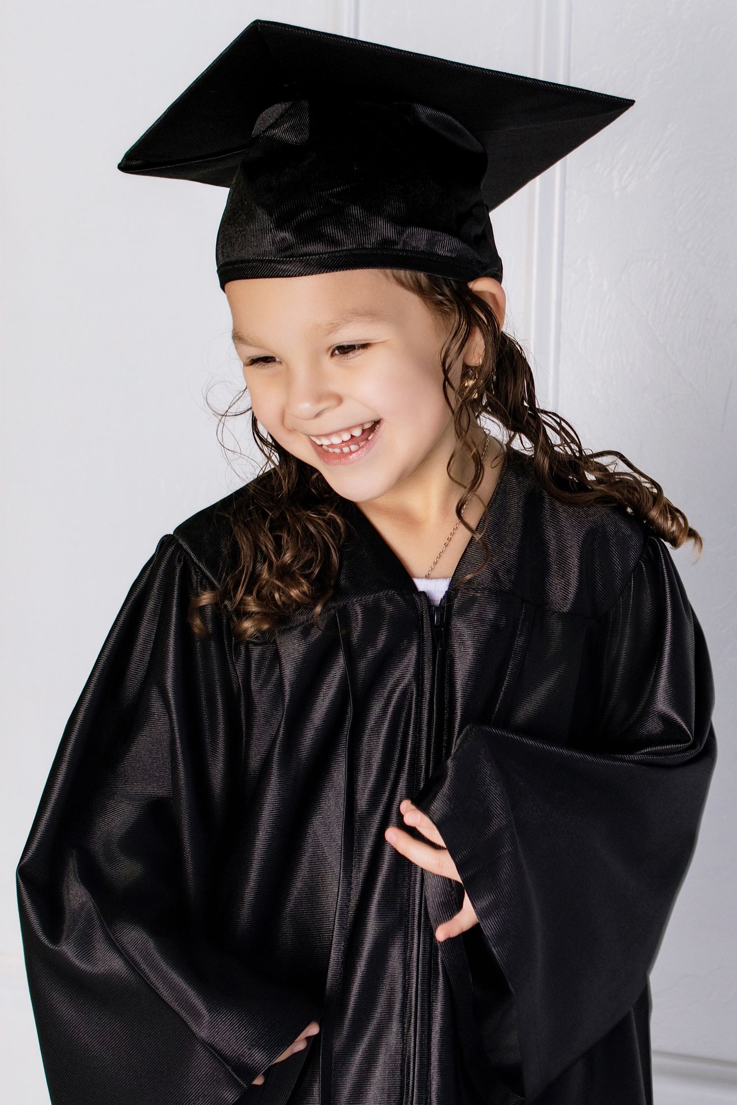 Smiling young girl in black graduation cap and gown posing against white background.