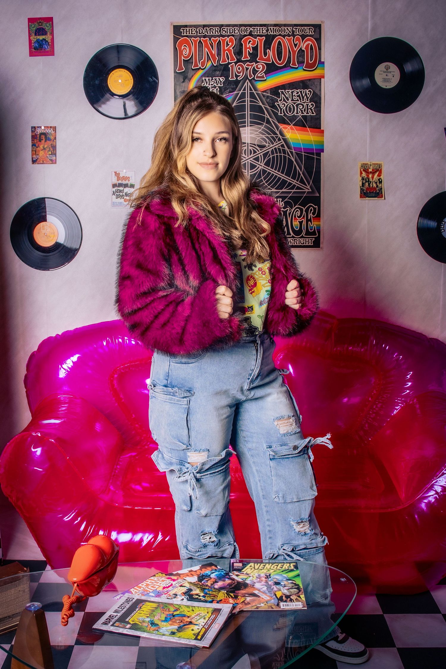 Teen girl in purple fur jacket and ripped jeans posing by pink couch with Pink Floyd poster and vinyl records.