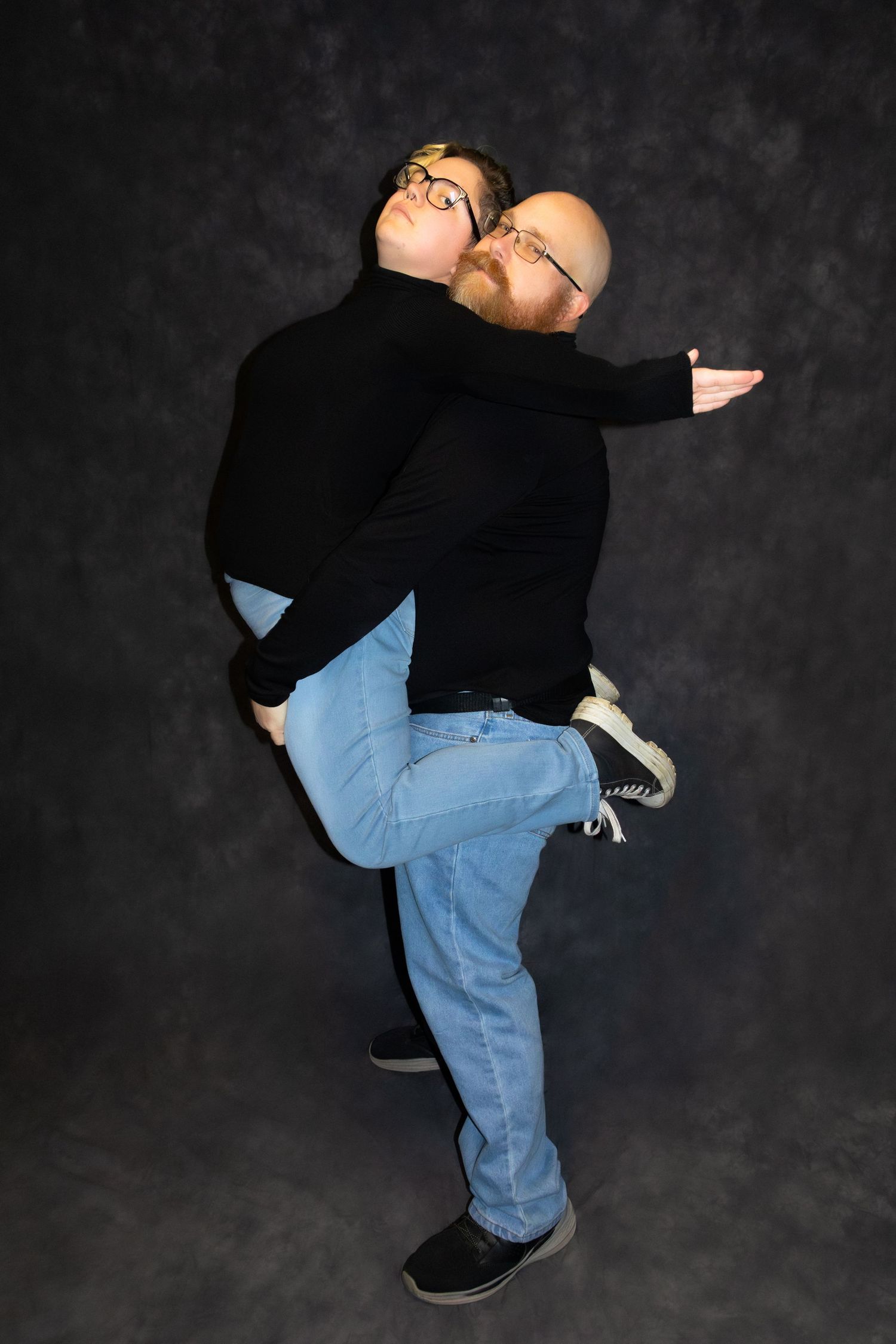 Two men in matching black turtlenecks and jeans pose playfully, one lifting the other in a studio setting.