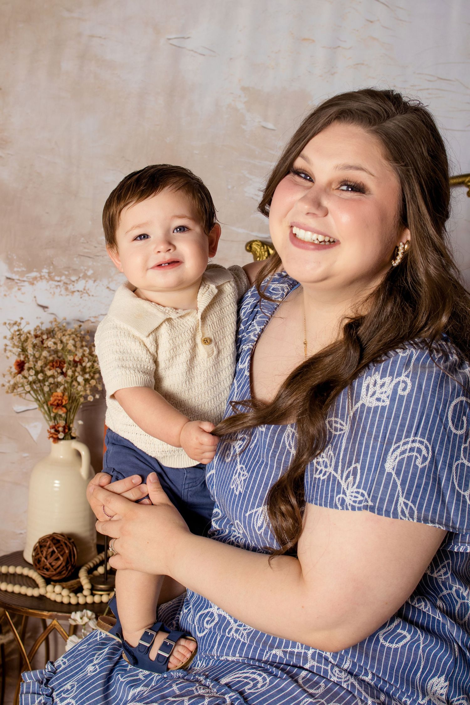 A smiling mother in a blue floral dress holds her happy baby boy in a cozy studio portrait setting.