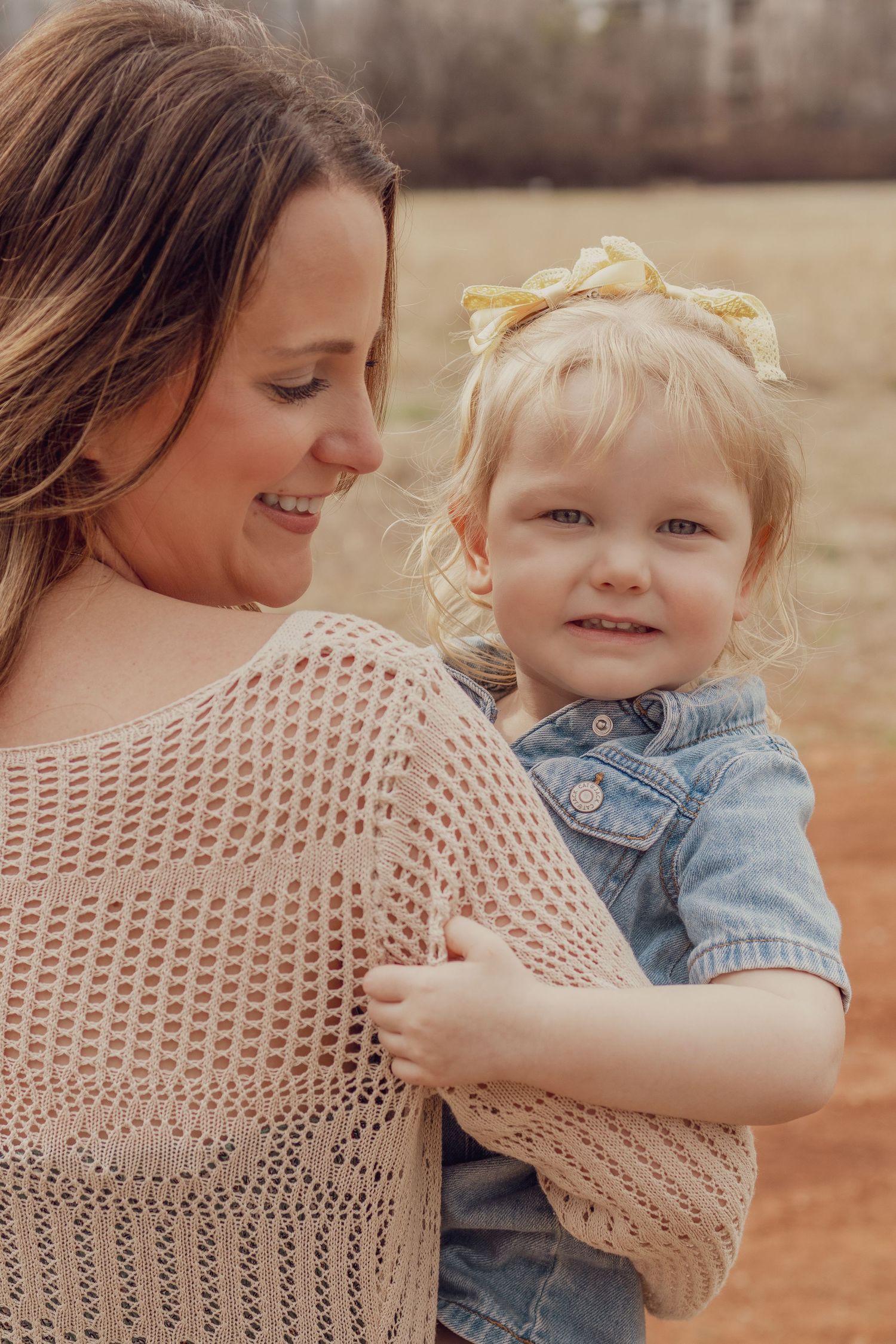 A tender moment as a mother holds her blonde toddler girl with a yellow bow in a natural outdoor setting.