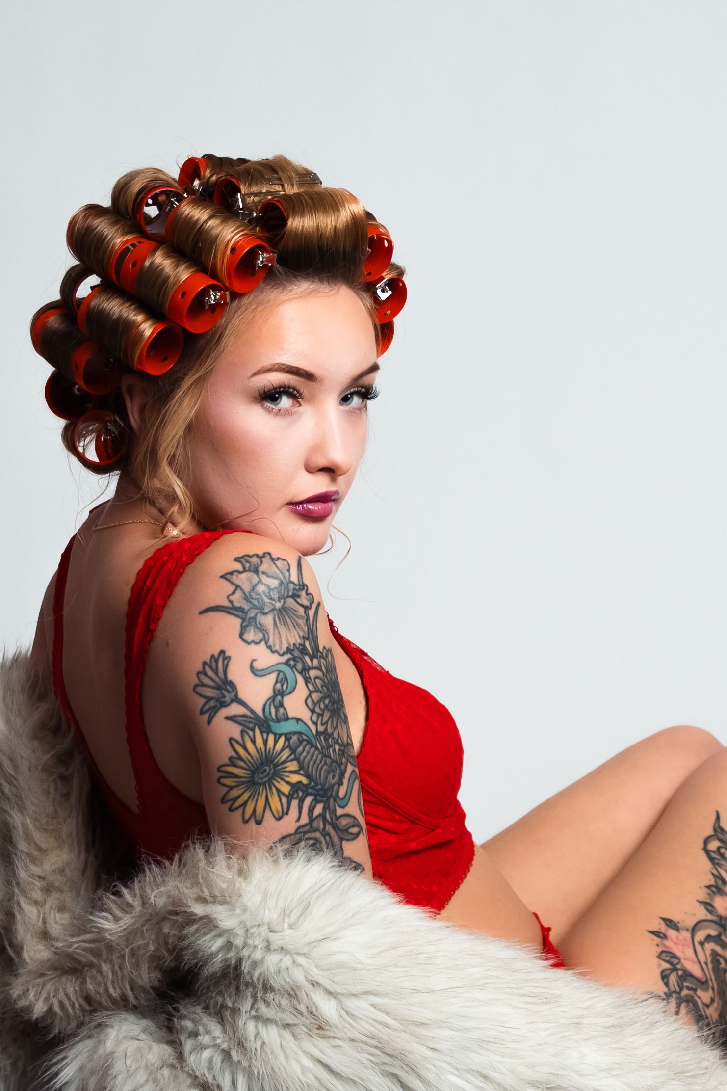 Woman in red lingerie with hair rollers and floral arm tattoo poses on fur, studio shot.