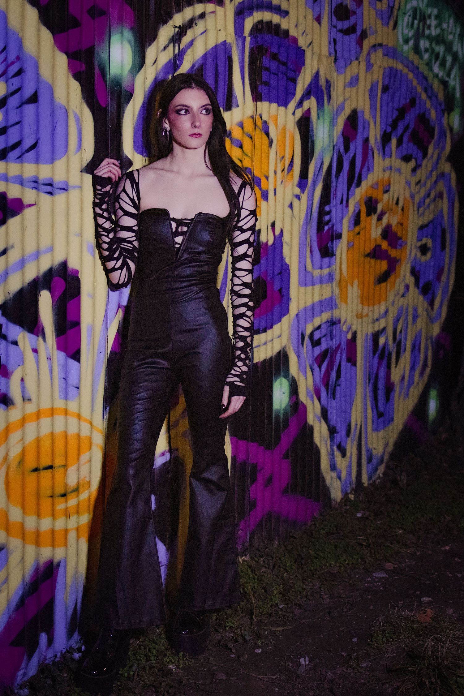 Woman in black leather jumpsuit with lace sleeves posing against colorful graffiti wall at night.