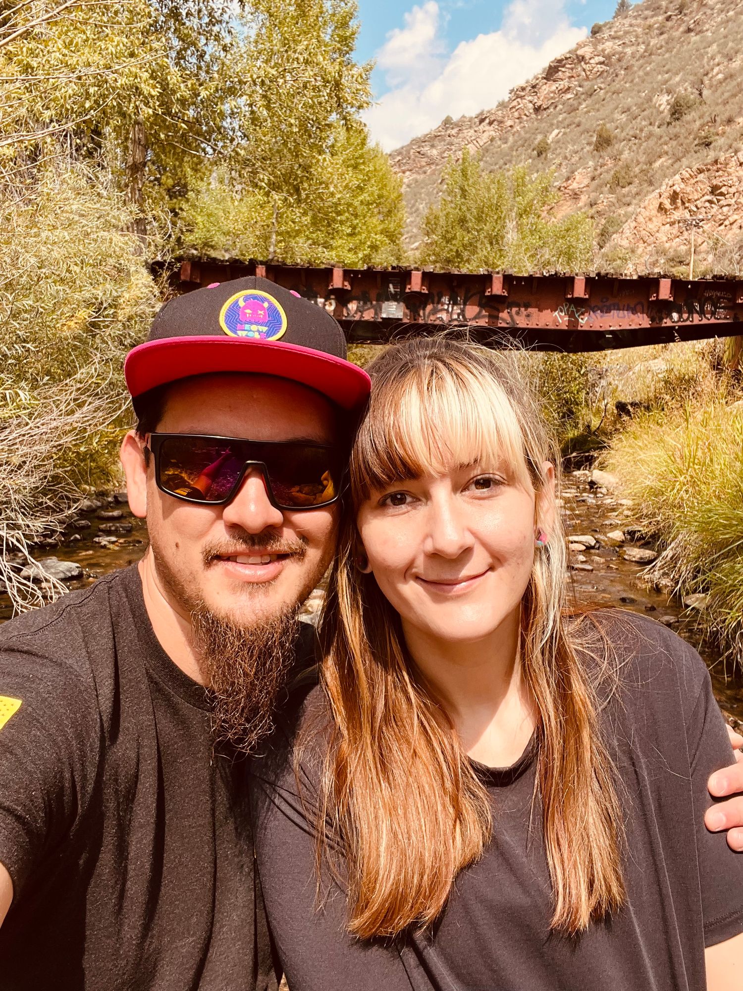 A couple smiles for a selfie near a rustic iron bridge over a creek, surrounded by lush trees and red rock cliffs.